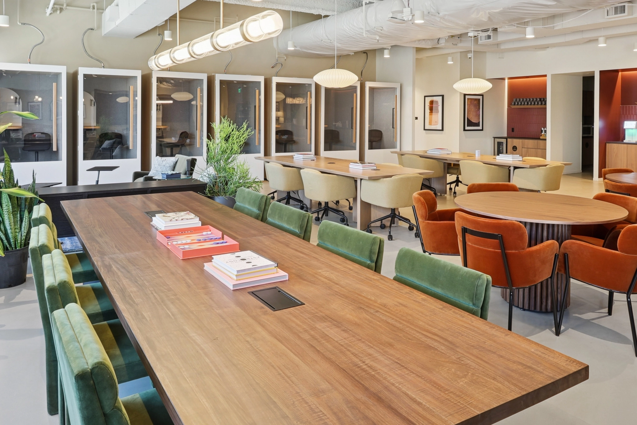 Modern coworking office space in The Woodlands with long wooden tables, various chairs, potted plants, and shelves with books, featuring a minimalist design and bright lighting.