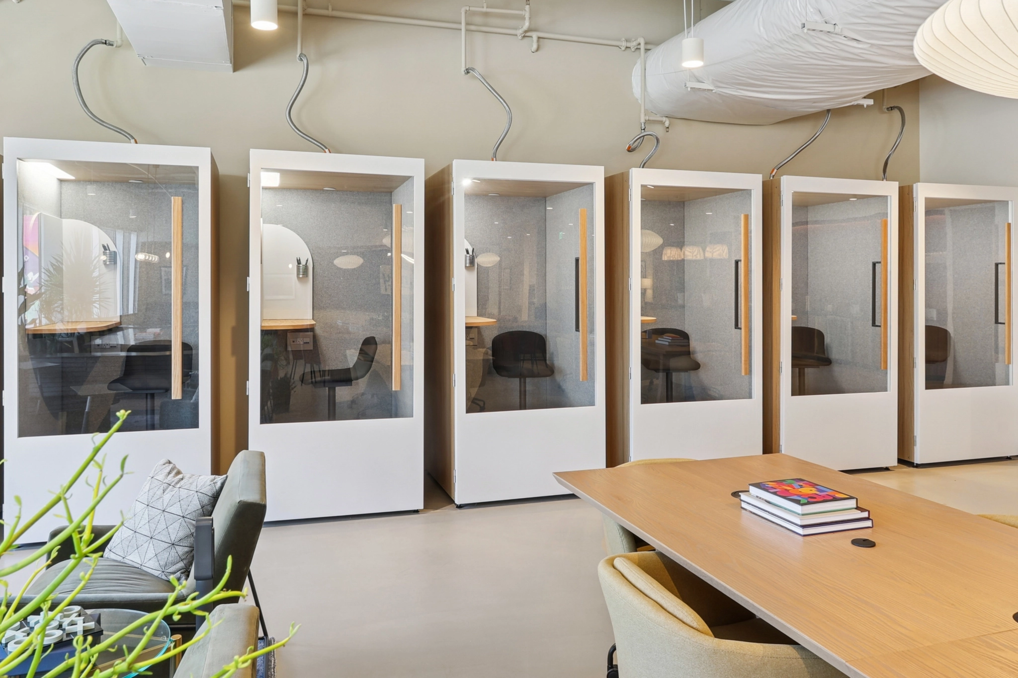 Six soundproof office phone booths with glass doors are lined up against a wall in this modern office; a table with books and chairs is in the foreground, creating a perfect setting for meetings in The Woodlands.