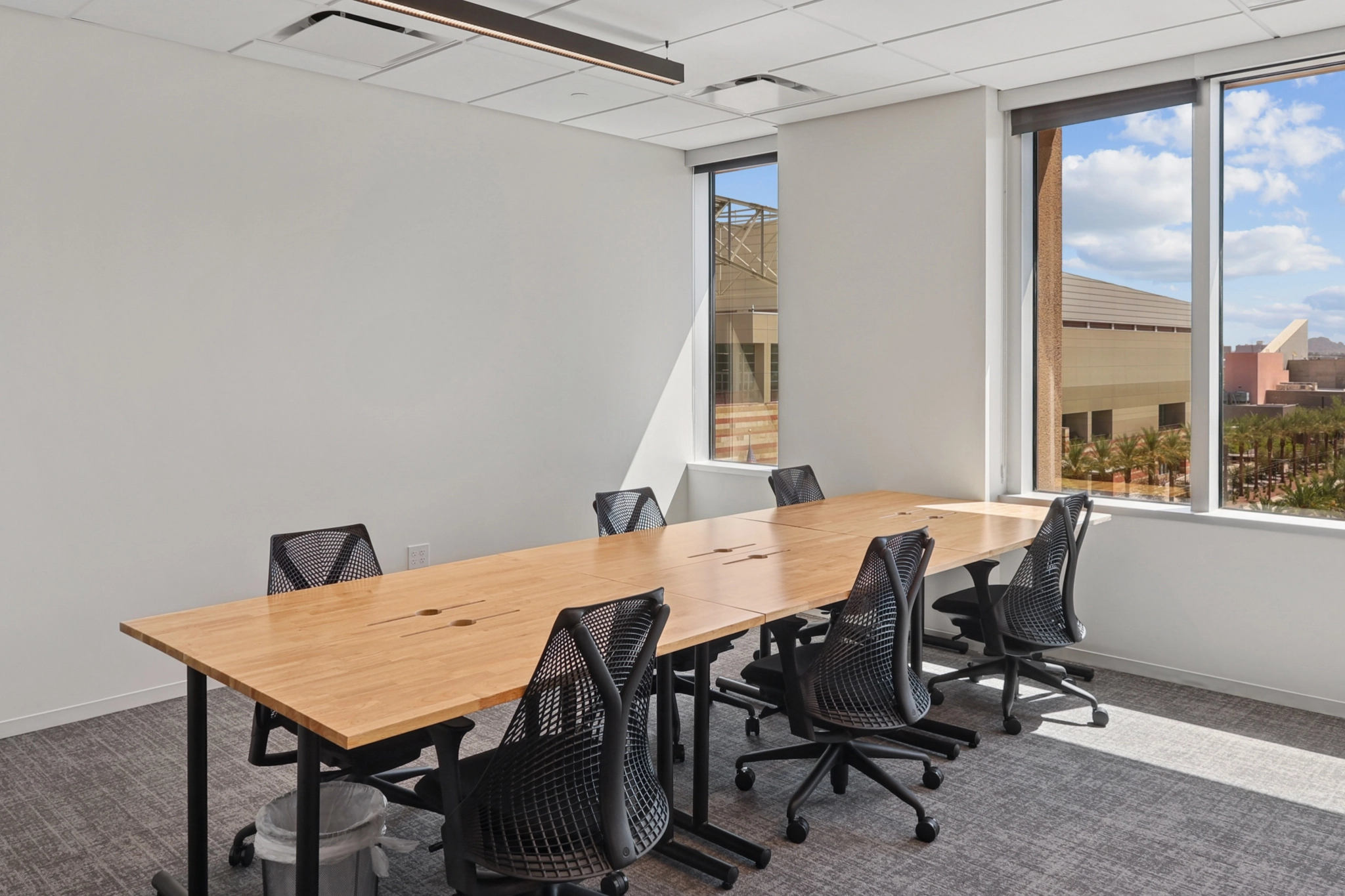 A modern coworking conference room with a wooden table, six black office chairs, and large windows overlooking buildings and a courtyard in The Woodlands.