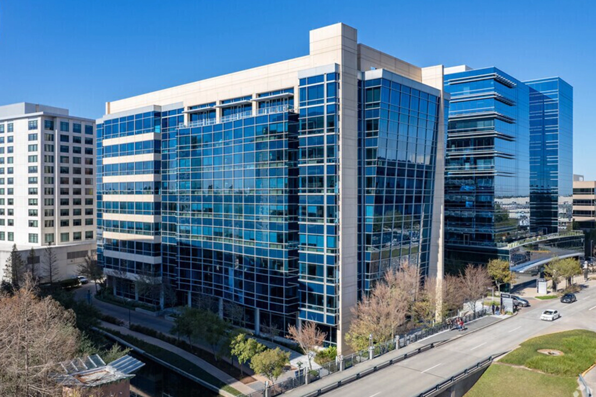 Modern office buildings with glass exteriors stand along a tree-lined street and small canal in The Woodlands, all beneath a clear blue sky.