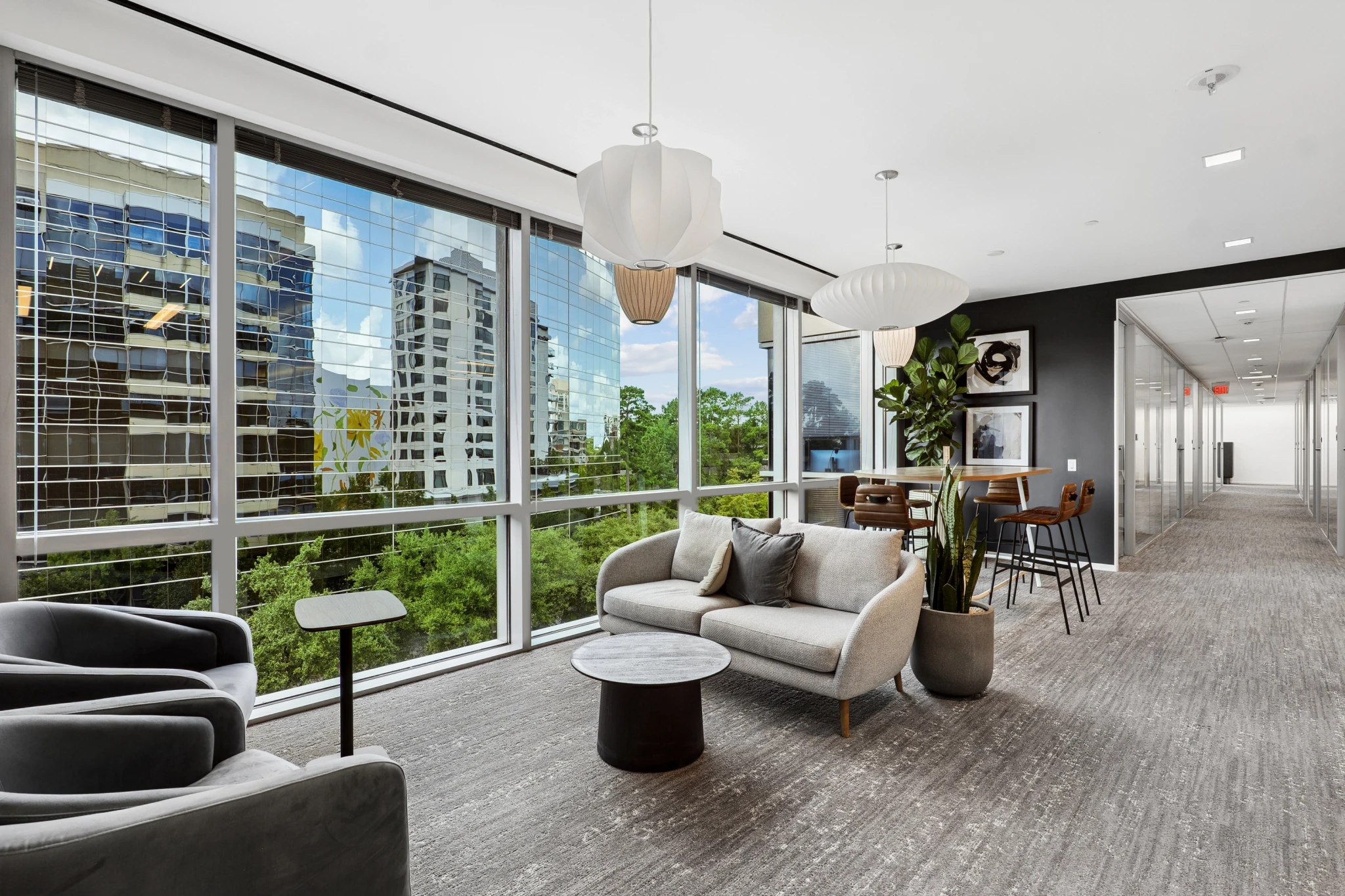 Moderner Bürosalon mit großen Fensterfronten, grauen Stühlen, runden Tischen, Zimmerpflanzen und Hängeleuchten; im Hintergrund sind die Gebäude der Stadt und die Bäume von The Woodlands zu sehen. Ein Flur führt zu einem Besprechungsraum und einem Essbereich, die sich im Hintergrund befinden.