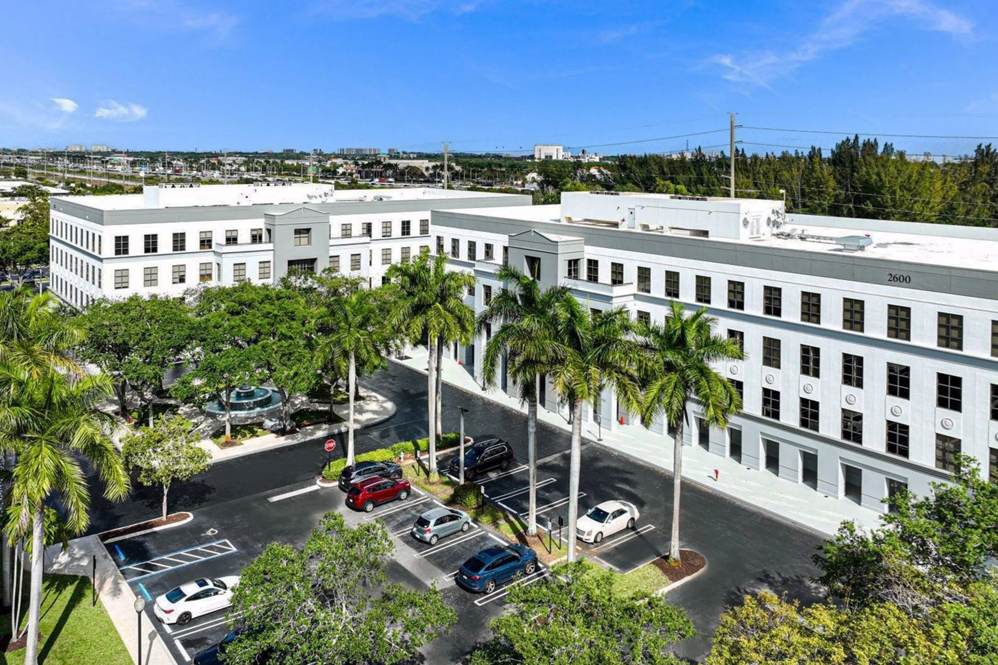 Aerial view of a white, three-story office building with palm trees, a parking lot, and a small fountain in front under a clear blue sky, offering modern office spaces and meeting rooms.
