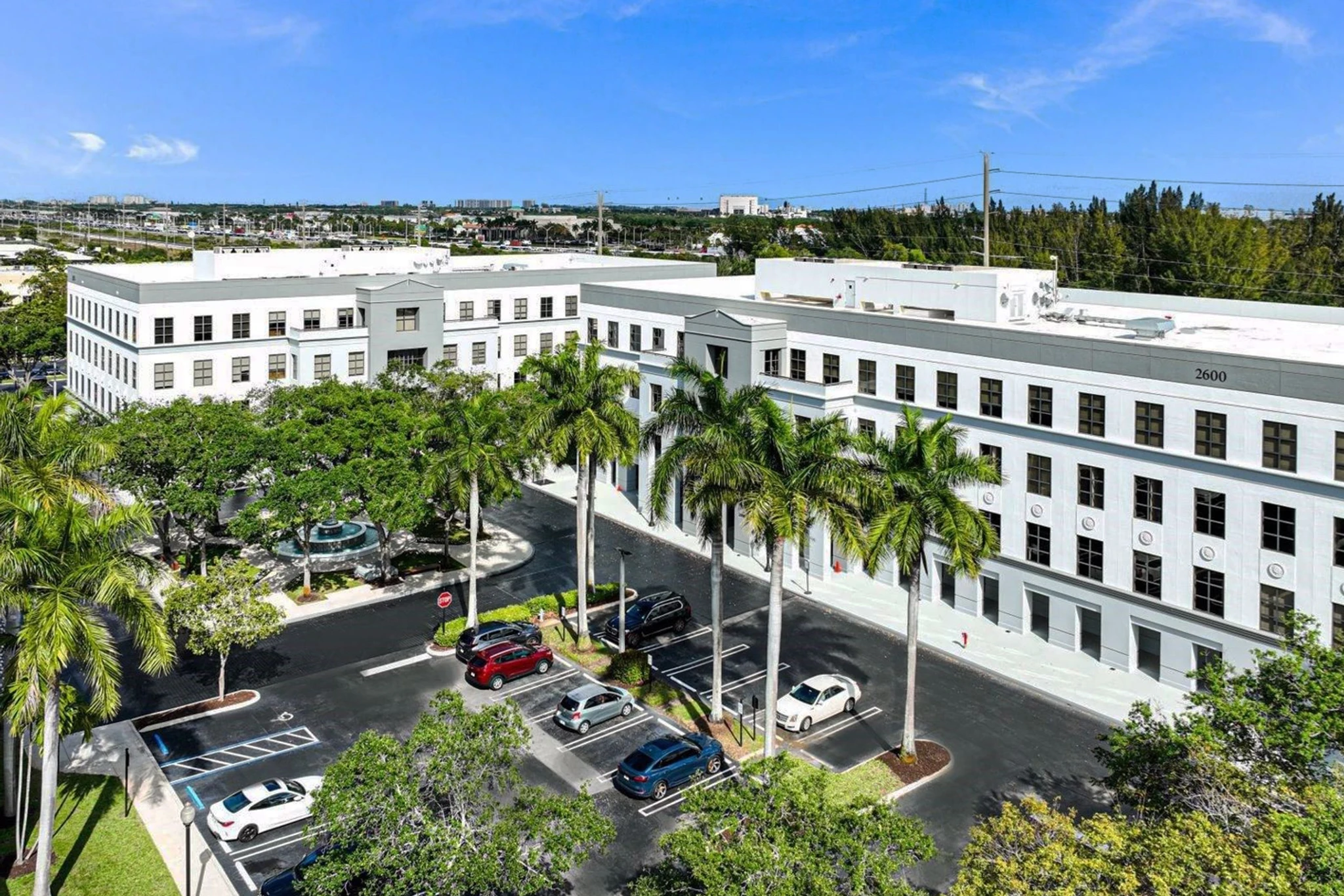Aerial view of a white, three-story office building with palm trees, a parking lot, and a small fountain in front under a clear blue sky, offering modern office spaces and meeting rooms.