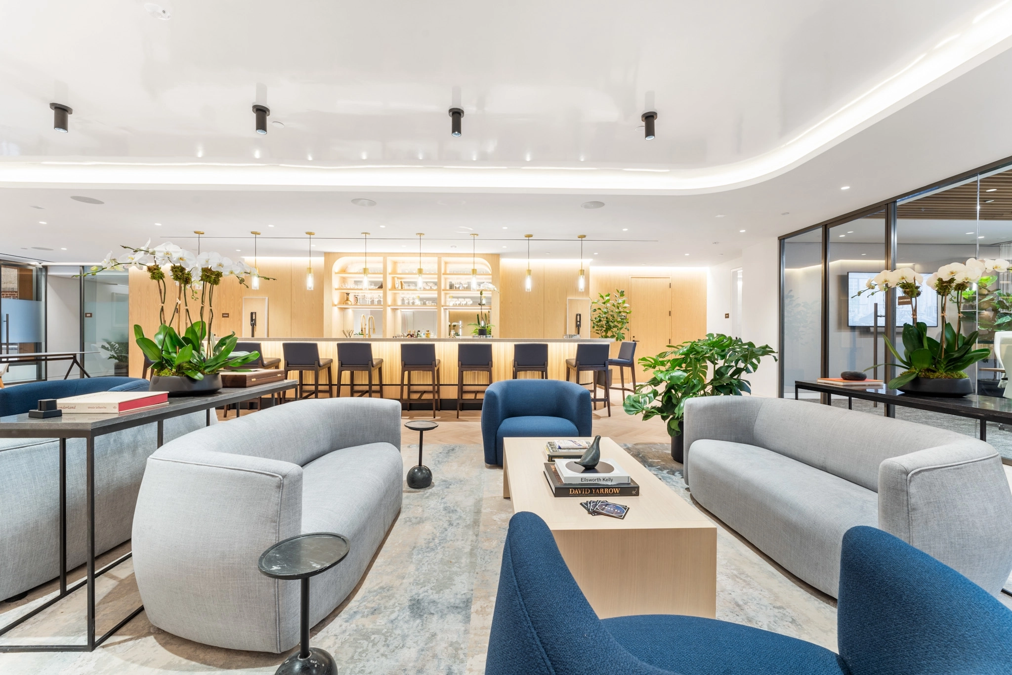 Modern office lounge area with curved sofas, armchairs, coffee tables, plants, and a bar counter with stools in the background under bright lighting—ideal for informal meetings or relaxing breaks in New York.