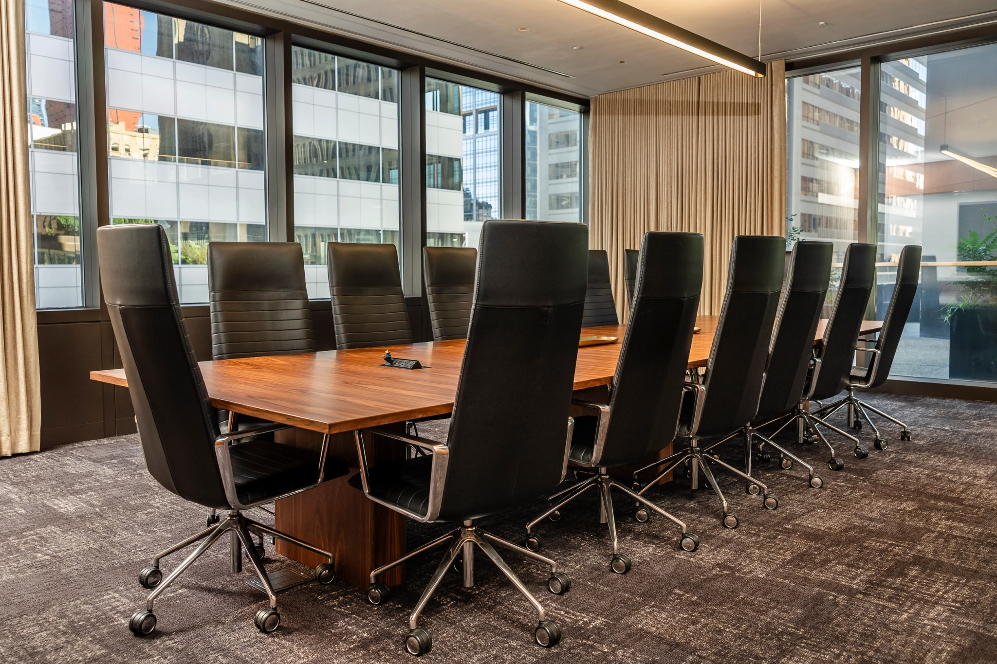 A modern meeting room with a large wooden table and twelve black office chairs, set against floor-to-ceiling windows showcasing Seattle’s skyline and tall office buildings outside.