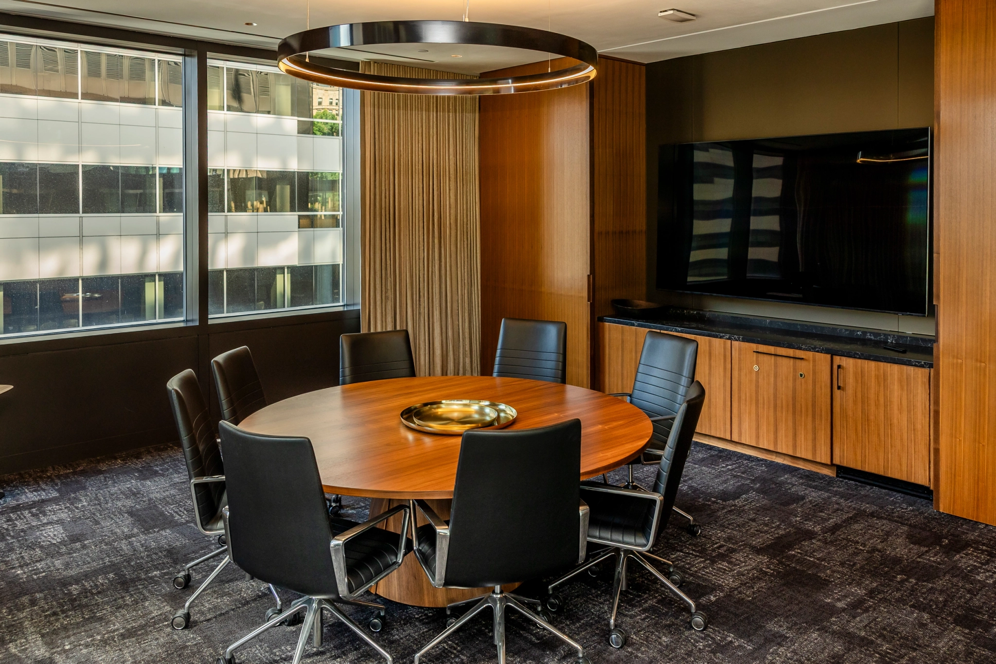 A modern meeting room with a round wooden table, six black chairs, a large wall-mounted screen, and windows overlooking Seattle office buildings.