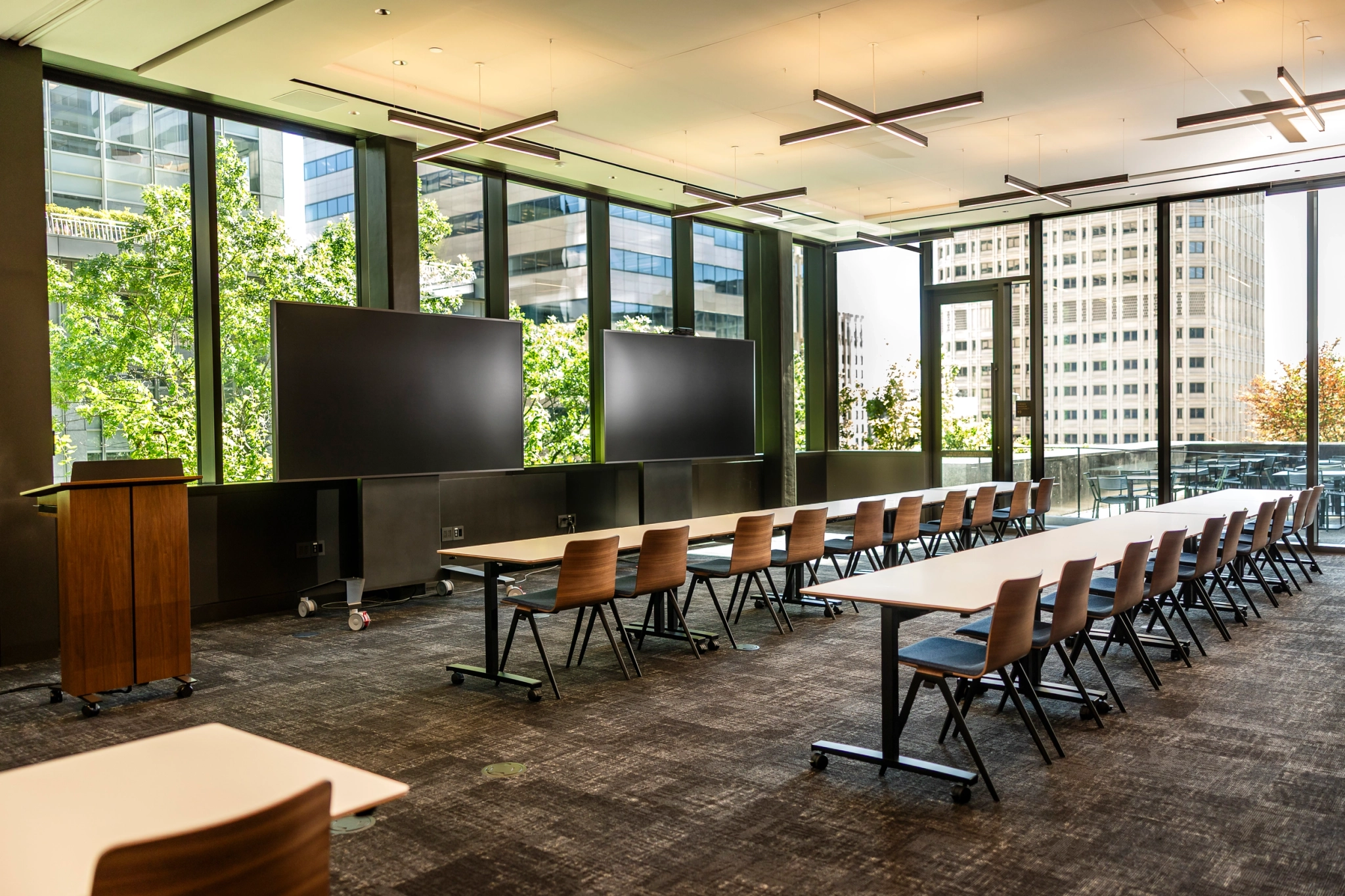 Modern classroom designed like a coworking space, featuring rows of empty desks and chairs, large windows overlooking city buildings, and two large screens at the front near a wooden podium.