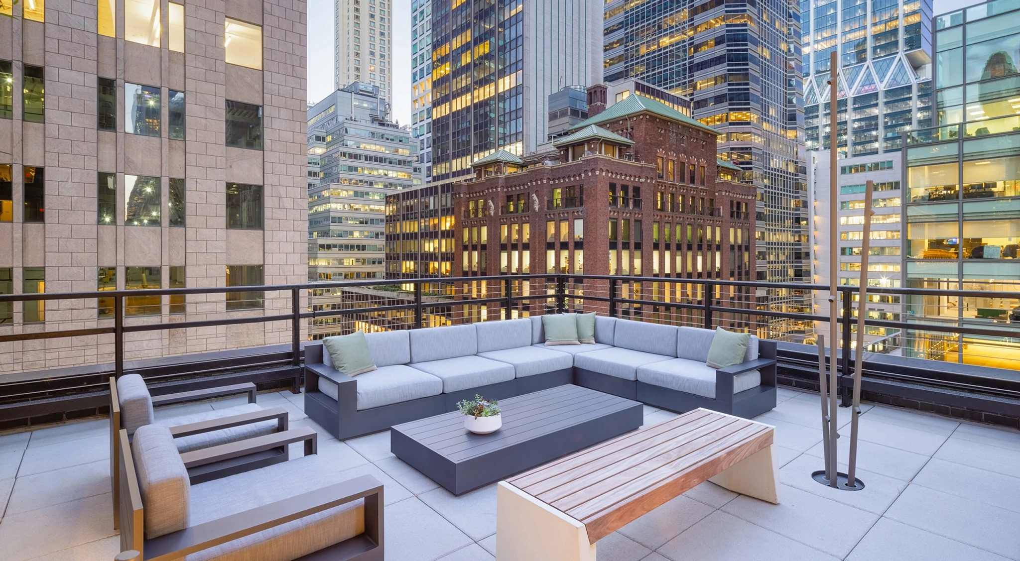 Rooftop patio with modern gray seating, a wooden bench, small tables, and potted plants, perfect for coworking and relaxation amid tall New York office buildings at dusk.