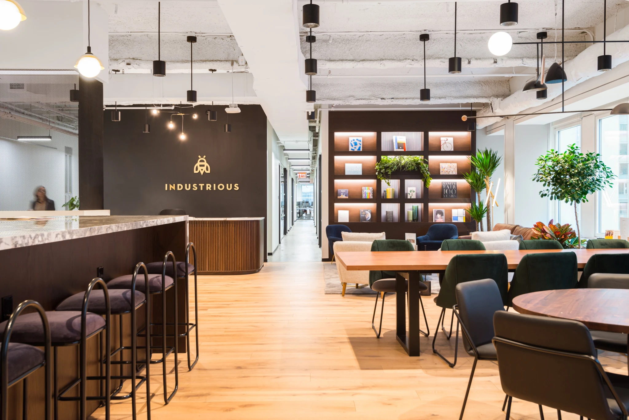 Modern coworking space in New York with bar seating, tables, greenery, bookshelves, and large windows, featuring the Industrious logo on a black accent wall—a vibrant workspace designed for productivity.