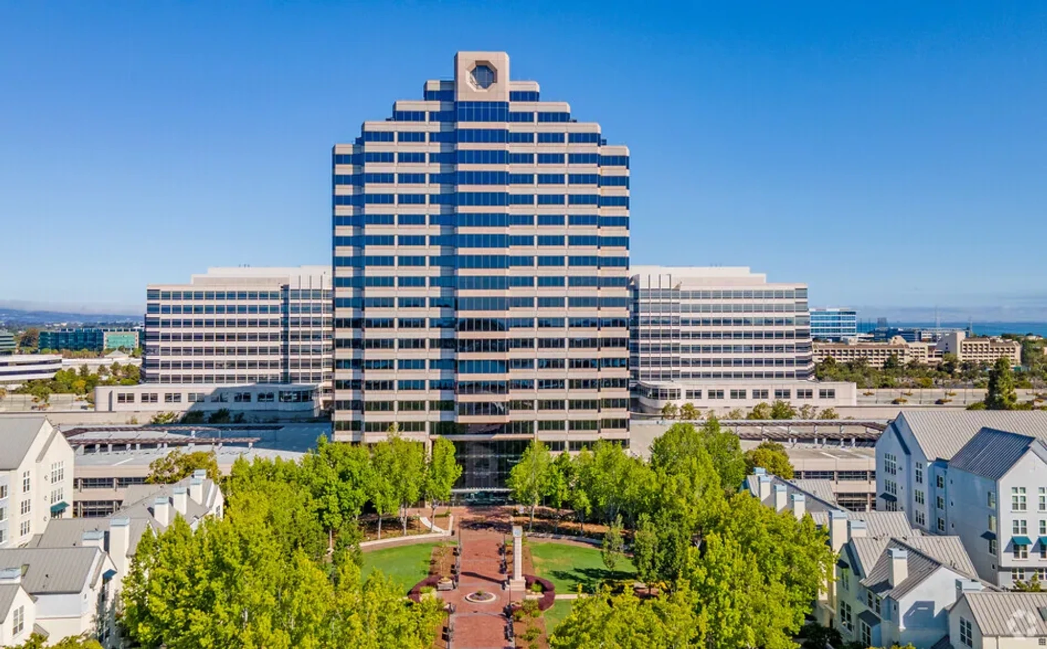 A large, modern office building with a stepped design stands behind a landscaped park with trees and pathways in Foster City on a clear, sunny day.