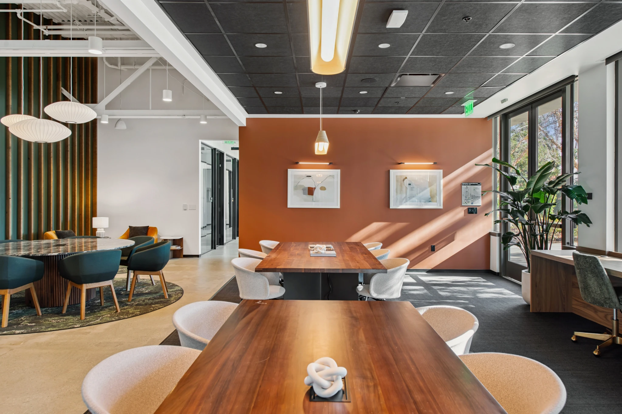 Modern office space in Coral Gables with wooden tables, white chairs, pendant lights, a green accent wall, framed art, indoor plants, and large windows letting in natural light—perfect for an inviting workspace or meeting room.