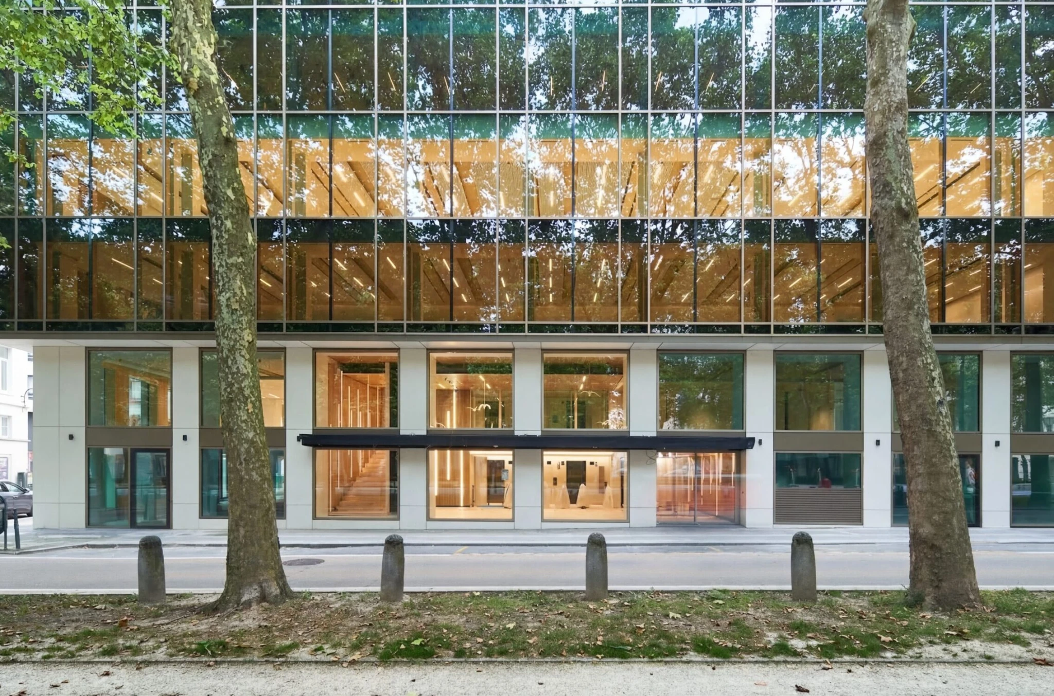 Ein modernes Bürogebäude aus Glas mit großen Fensterfronten, Holzakzenten im Innenbereich und dem Spiegelbild der Bäume an der Fassade; es beherbergt Coworking-Bereiche und Besprechungsräume, während gegenüber die Straße von Bäumen und Pollern gesäumt ist.