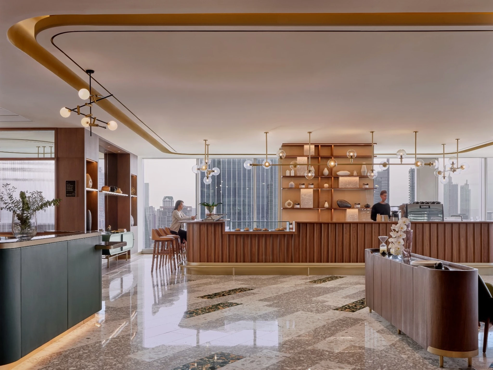 Modern hotel lobby with marble floors, wooden reception desk, decorative shelves, and floor-to-ceiling windows overlooking Bangkok's city skyscrapers. Two people stand near the counter, discussing a nearby meeting room workspace.