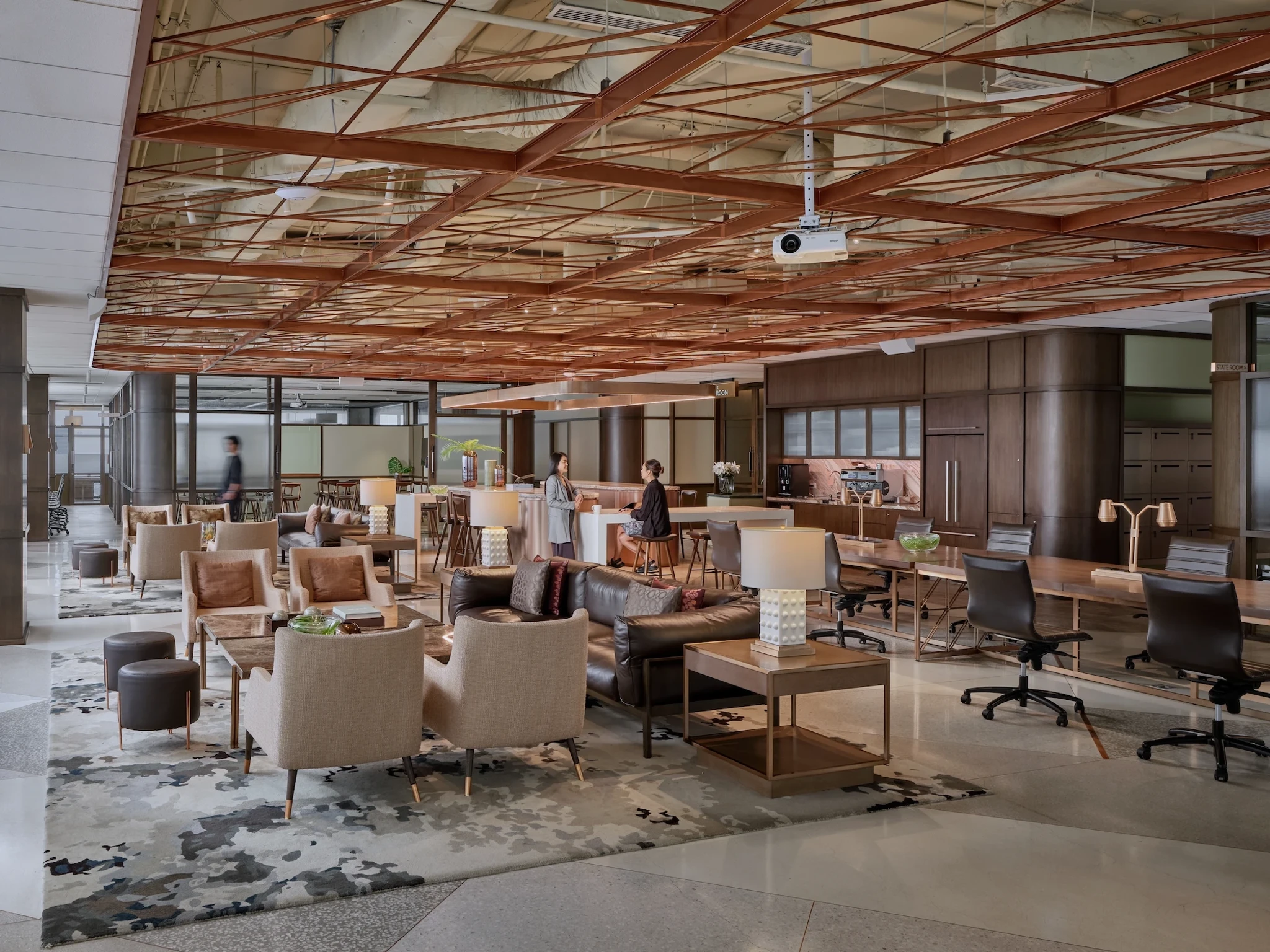 Modern office lounge with various seating areas, tables, a projector, and people working or talking. The space features a patterned ceiling and neutral-toned decor, creating an inviting coworking environment ideal for collaboration and productivity.