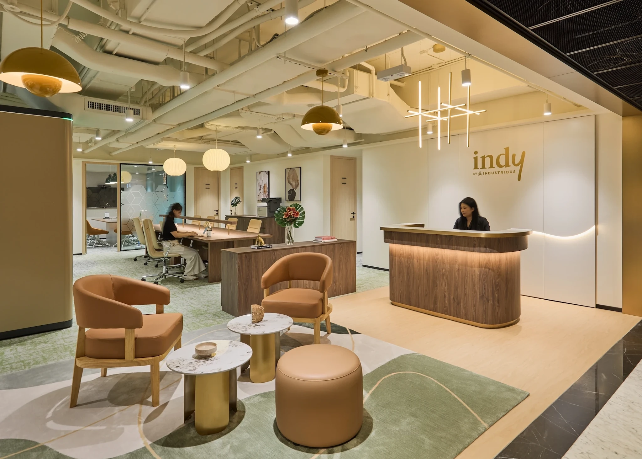 Modern office lobby with brown leather chairs, marble tables, pendant lights, a receptionist at a wooden desk, and "indy" sign on the wall