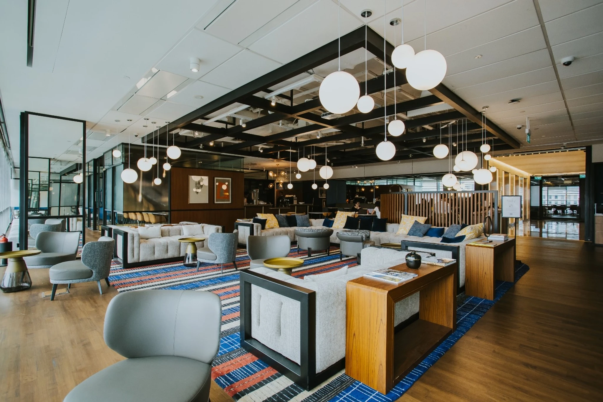 Modern lounge area with contemporary furniture, round hanging lights, patterned rugs, and a mix of sofas and chairs in a spacious, well-lit workspace perfect for informal meetings or relaxing breaks.