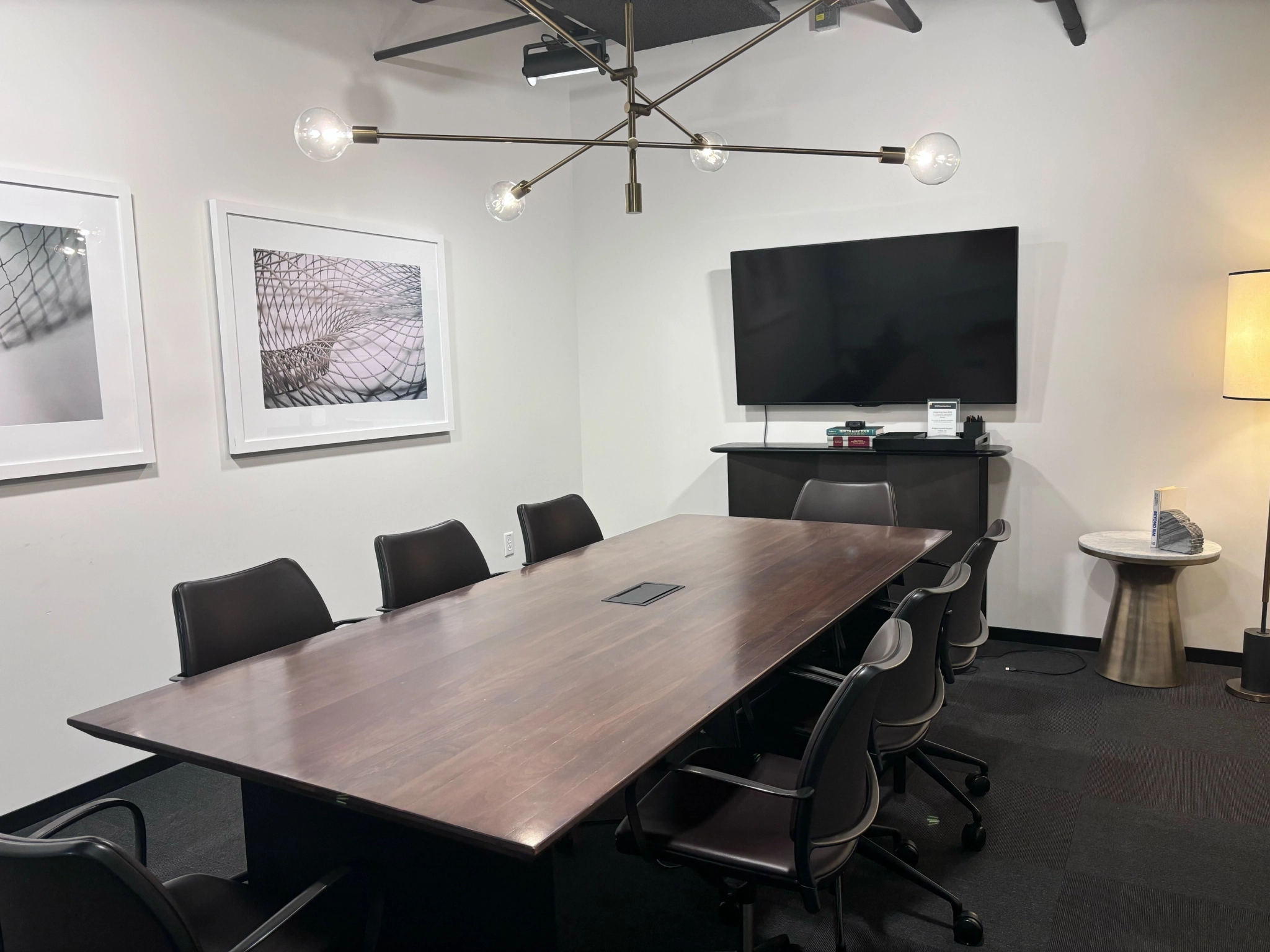 A modern Boston meeting room with a sleek wooden table, six chairs, a wall-mounted TV for presentations, abstract artworks adding a creative flair, and an elegant ceiling light fixture.