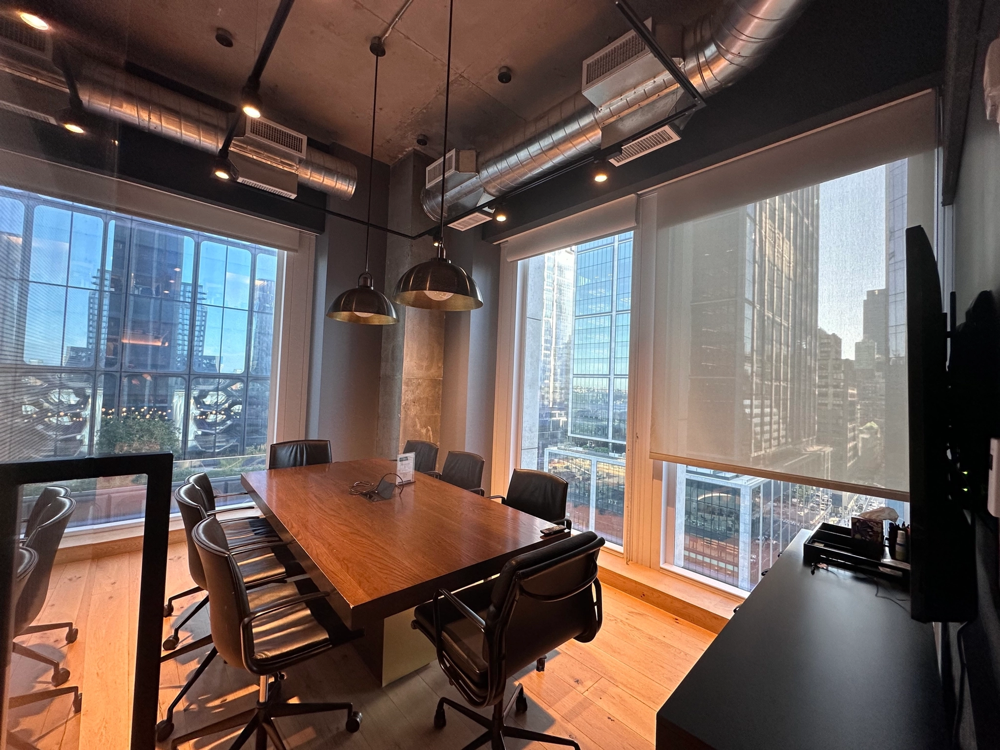 Modern meeting room with a large wooden table, black chairs, and overhead lighting. Large windows overlook the iconic New York cityscape with tall buildings, offering an inspiring backdrop for any gathering.