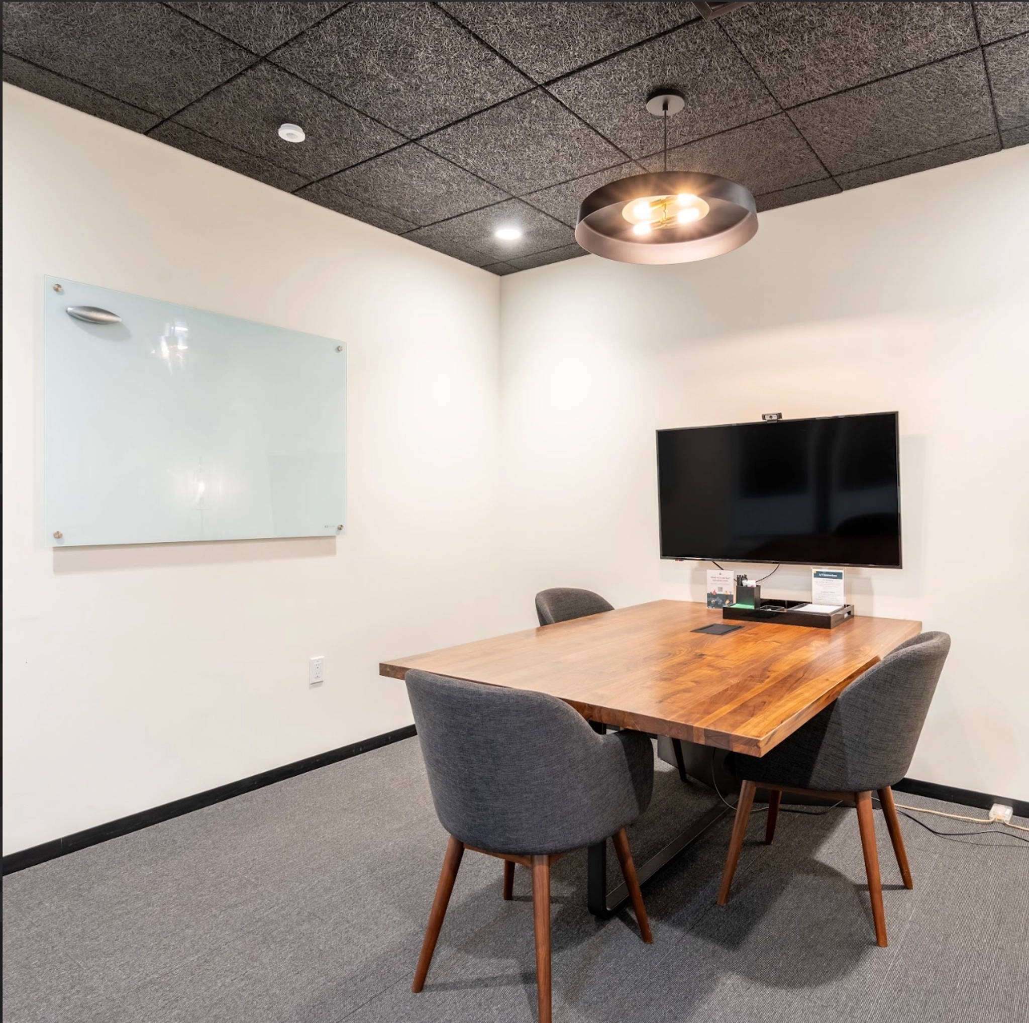 Moderne vergaderruimte met houten tafel, vier stoelen, televisie aan de muur en glazen whiteboard. Deze coworkingruimte heeft een zwart plafond met een enkele hanglamp, waardoor stijl en functionaliteit perfect worden gecombineerd.