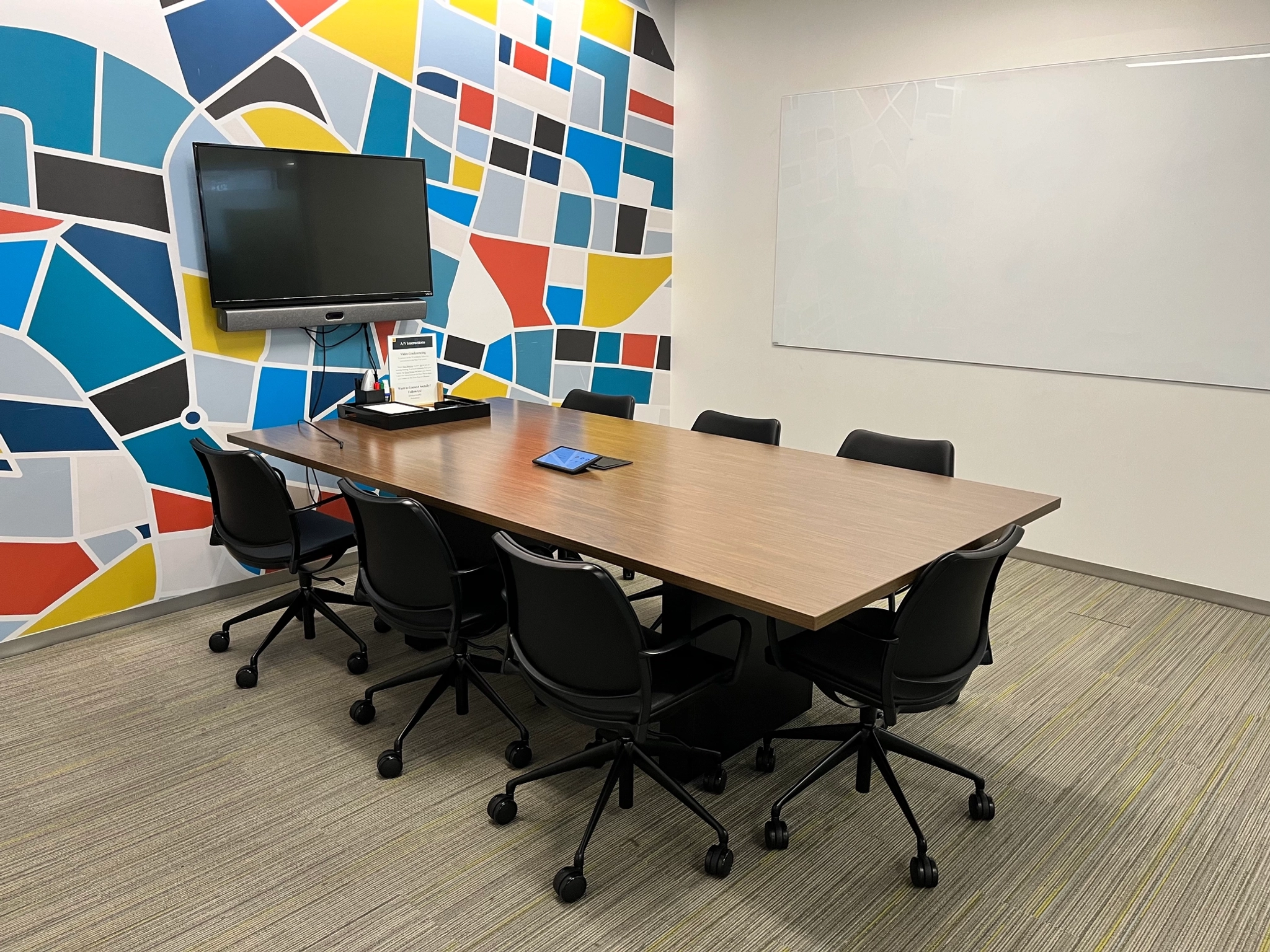 A meeting room with a rectangular table, six chairs, a wall-mounted TV, and a colorful geometric wall design. A whiteboard is visible on the adjacent wall, making it a versatile workspace for any team.