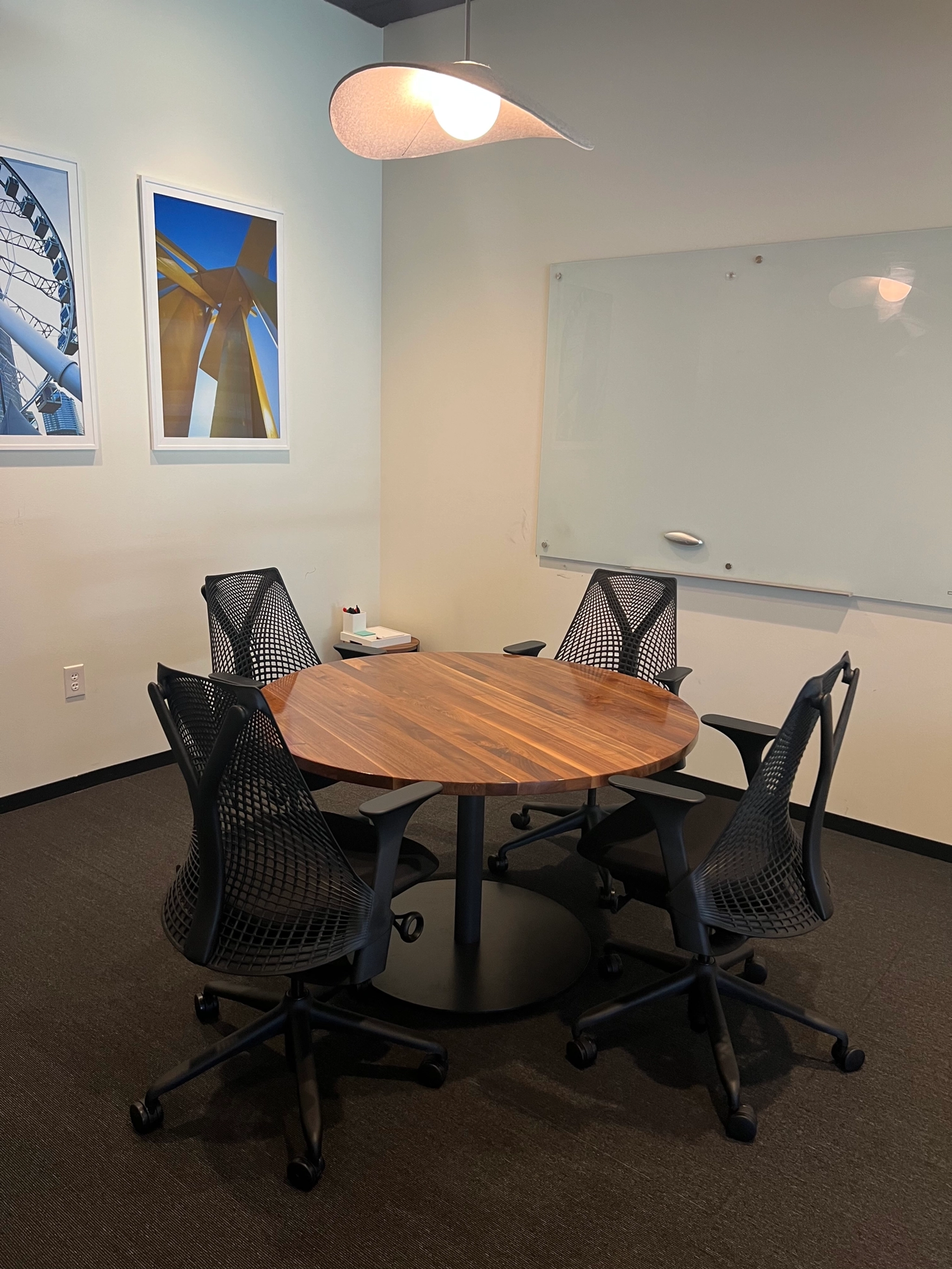 Une petite salle de réunion de coworking à Dallas avec une table ronde en bois, quatre chaises de bureau noires, un tableau blanc et deux photos architecturales encadrées au mur.