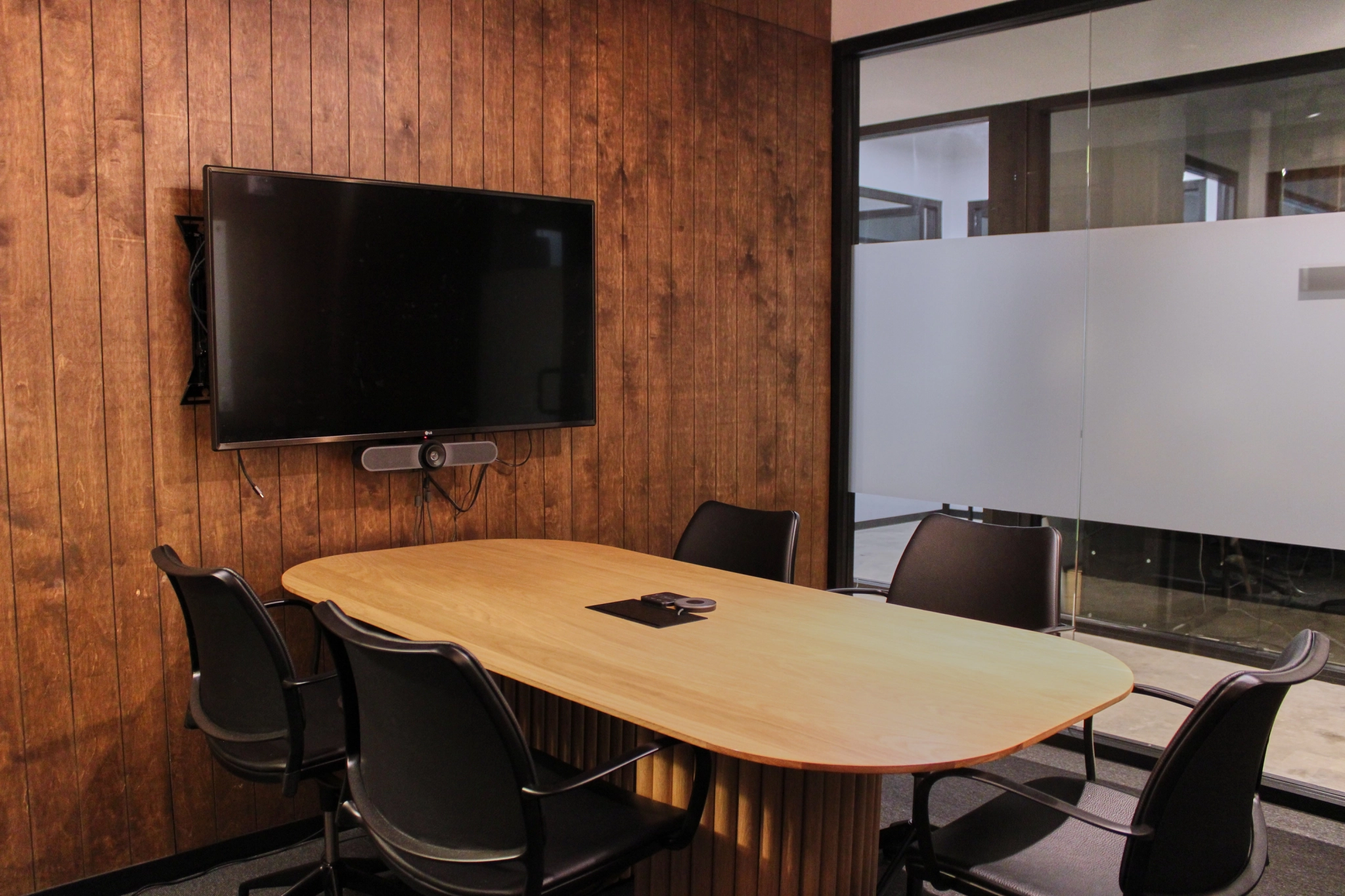 Een kleine moderne vergaderruimte met houten wanden, een aan de muur gemonteerde televisie, een tafel en vijf zwarte stoelen. Aan de rechterzijde is een glazen scheidingswand met matte panelen aanwezig. Deze goed ontworpen werkruimte biedt een ideale setting voor productieve kantoorgesprekken.