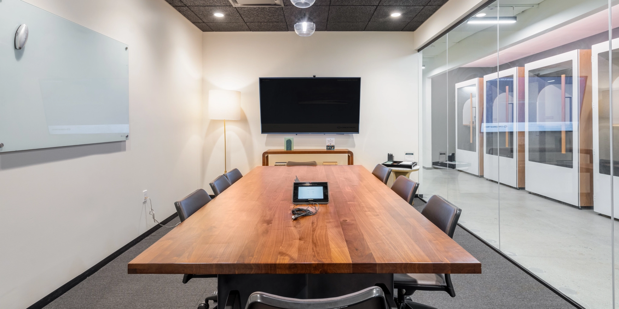 Un espace de travail moderne comprenant une table en bois, des chaises de bureau, un téléviseur mural, un tableau blanc et une cloison en verre, parfait pour tous les besoins d'une salle de réunion.