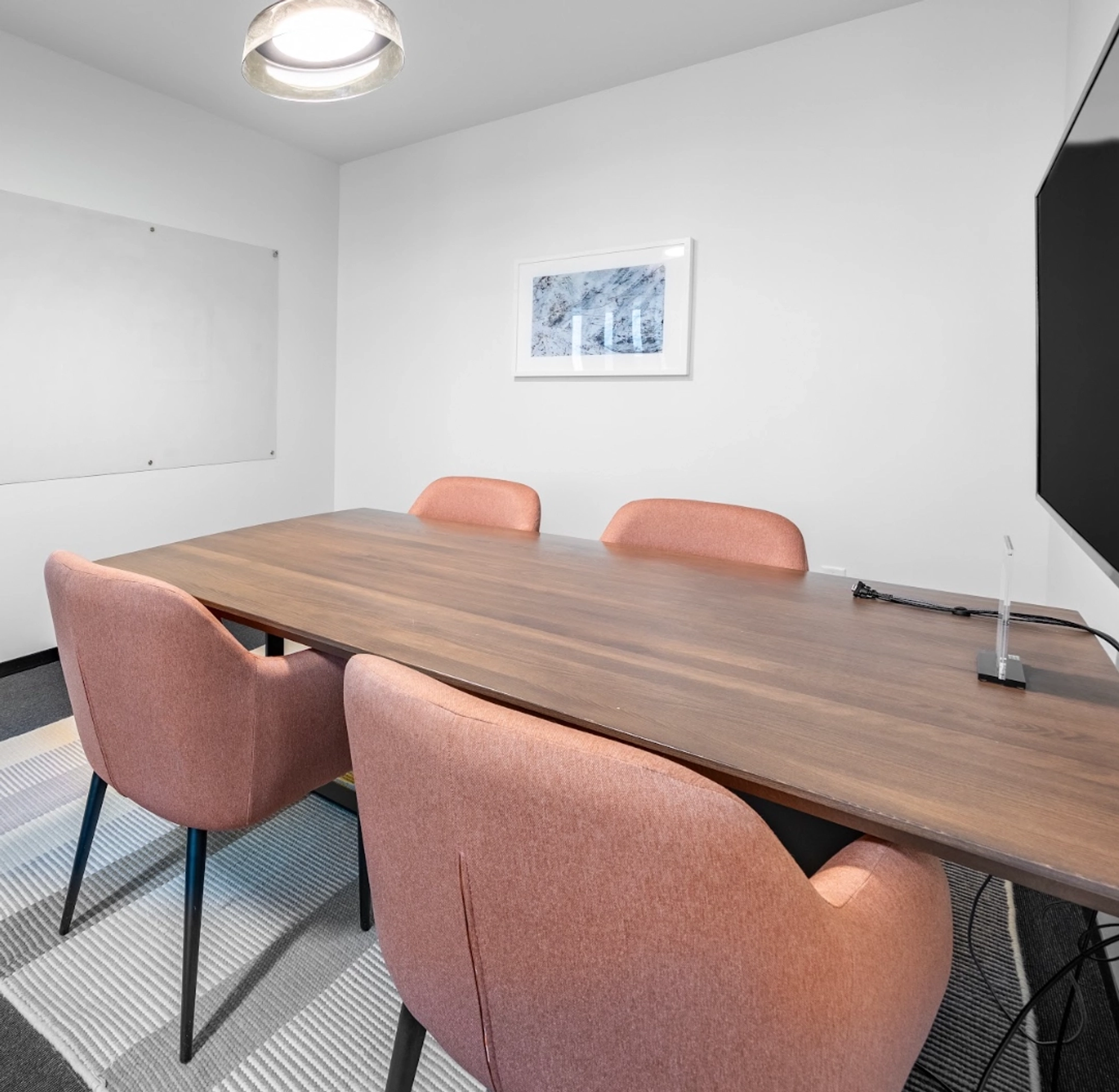 A small office meeting room with a wooden table, four pink chairs, a wall-mounted whiteboard, a framed picture, a ceiling light, and a wall-mounted TV.