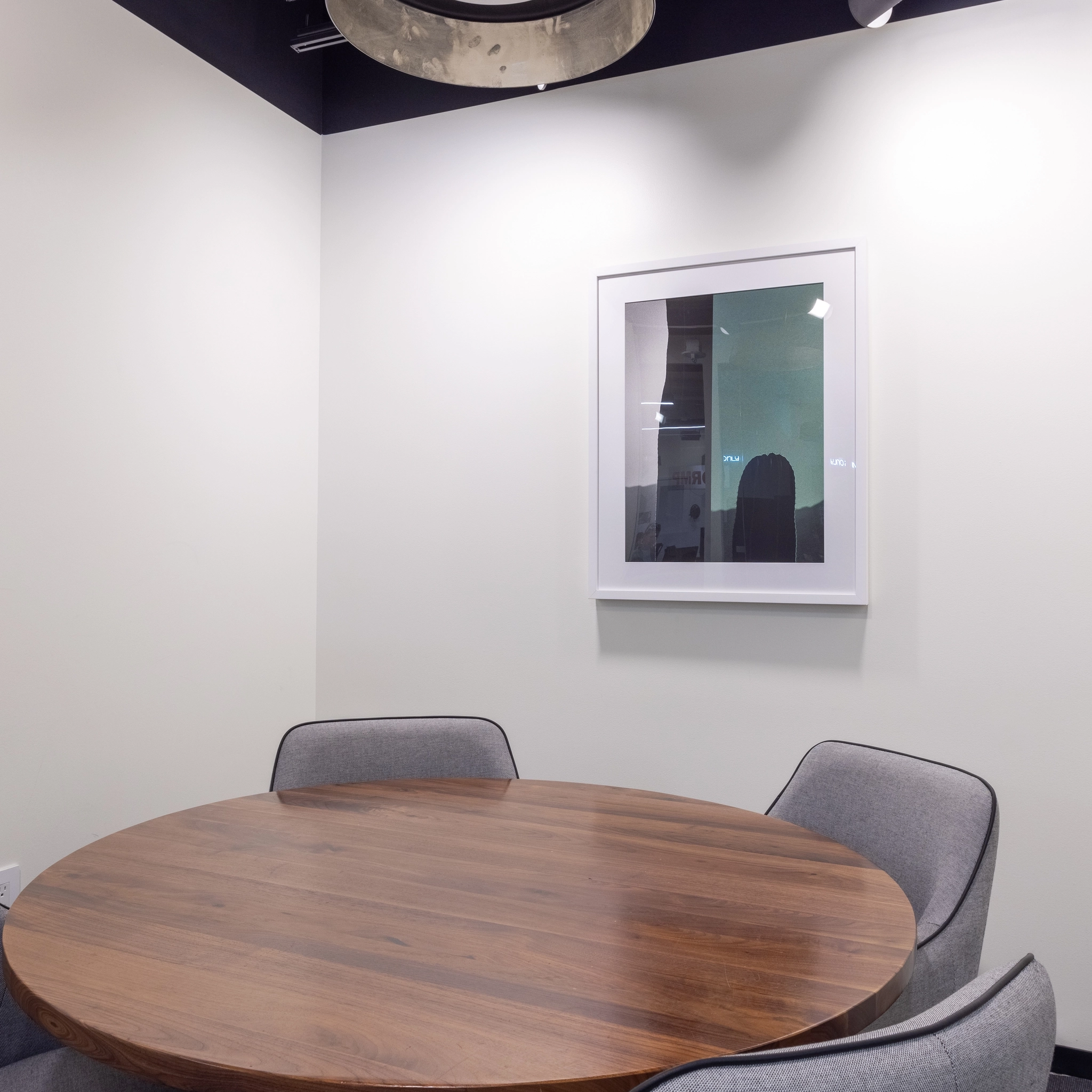 Une petite salle de conférence dans les bureaux de McLean comprend une table ronde en bois, quatre chaises grises et un tableau encadré sur le mur. La pièce est dotée de murs blancs et d'un plafonnier, ce qui la rend parfaite pour les séances de coworking.