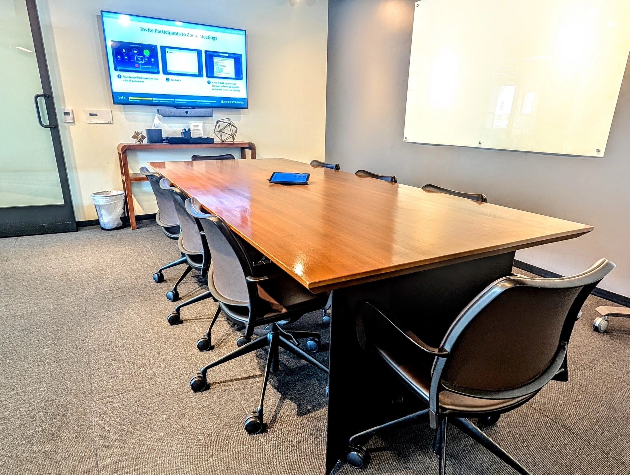 A meeting room with eight chairs around a wooden table, a tablet on the table, a large display screen on the wall, and a whiteboard—perfect for coworking or collaborative workspace needs.