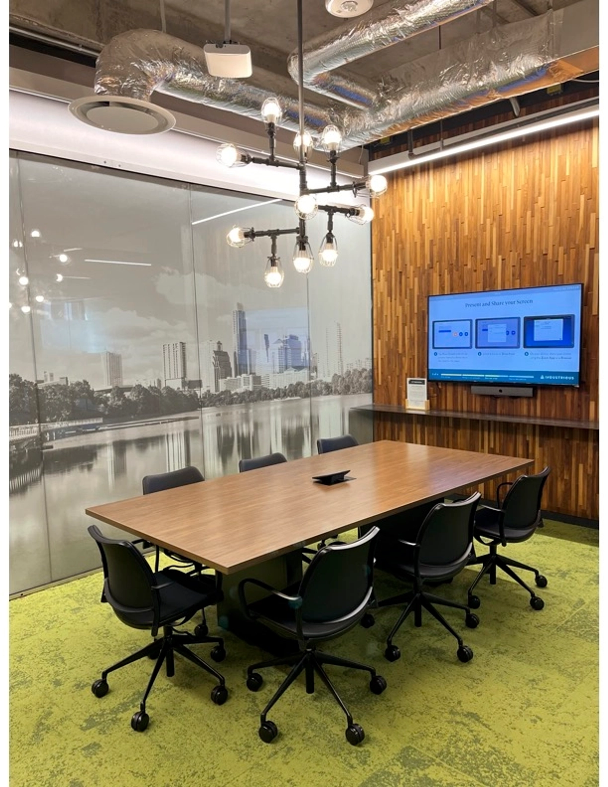 A modern meeting room with a wooden table, six chairs, a wall-mounted monitor displaying presentation slides, and a decorative light fixture. The background features a cityscape mural, making it an ideal workspace for collaboration.