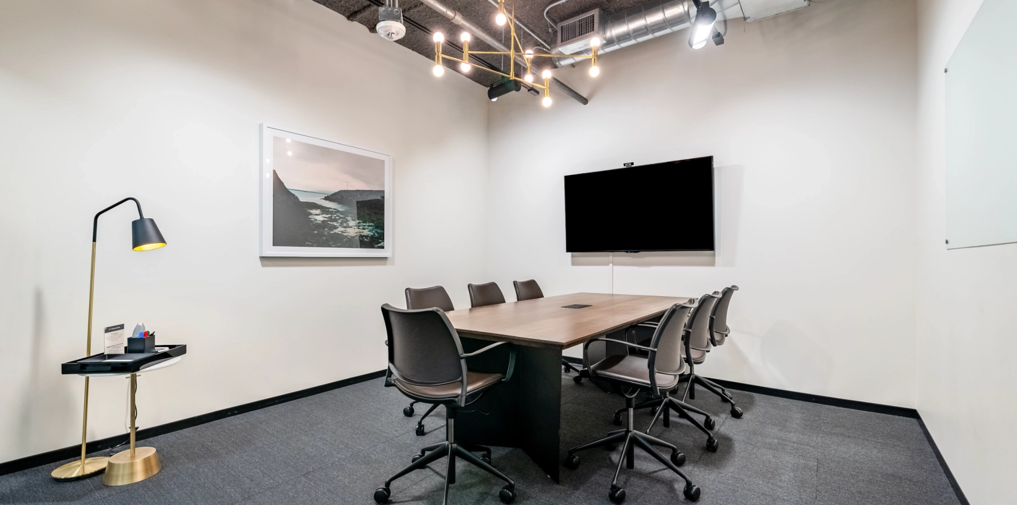 A modern coworking meeting room features a rectangular table, eight chairs, a wall-mounted TV, and a framed picture. A standing lamp and a small shelf with office supplies are tucked in the corner, enhancing this collaborative office space.