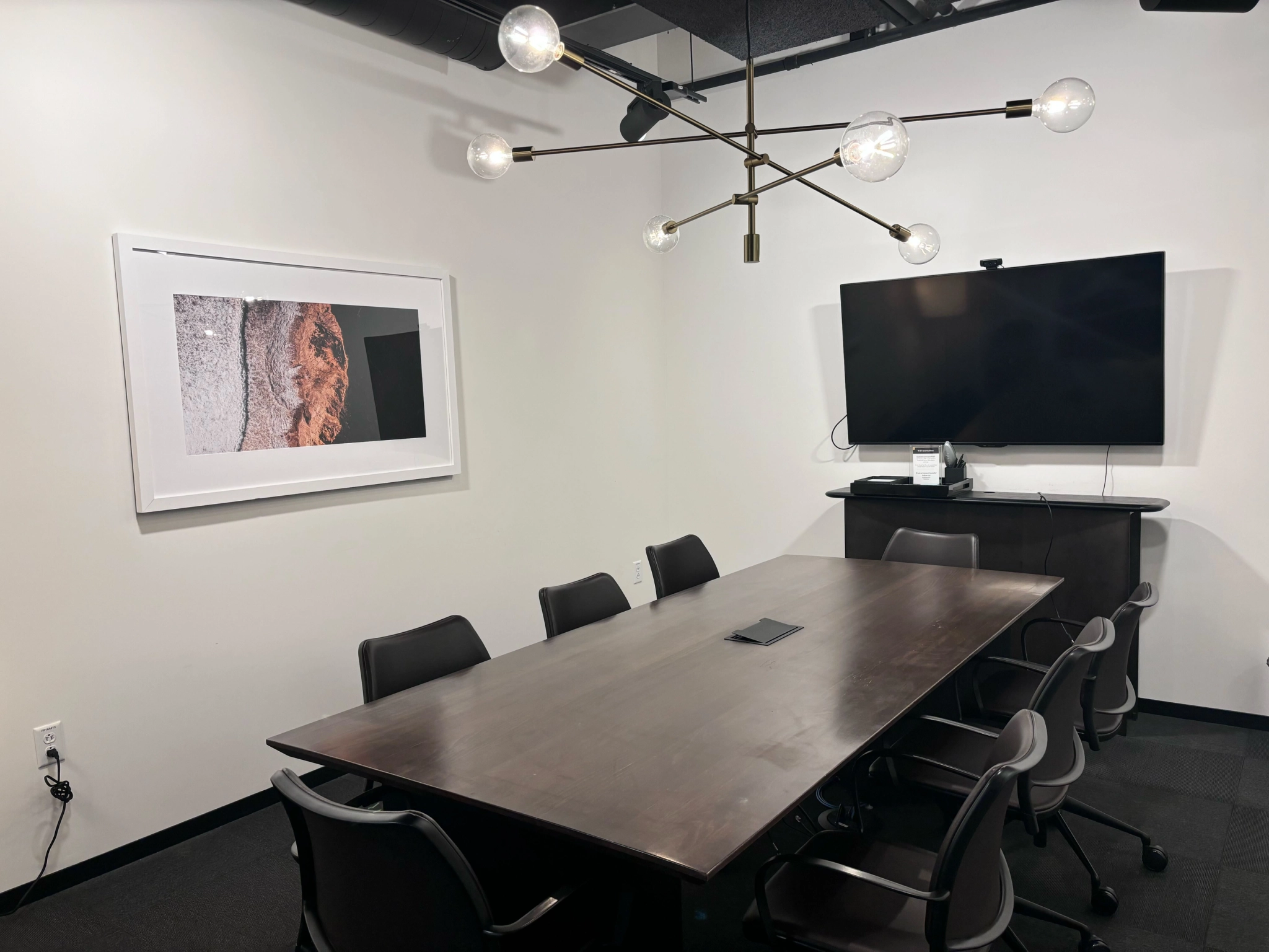 A modern conference room in a Boston office features a long wooden table, six chairs, a wall-mounted TV, and abstract artwork on white walls—perfect for coworking under the glow of a sleek chandelier.