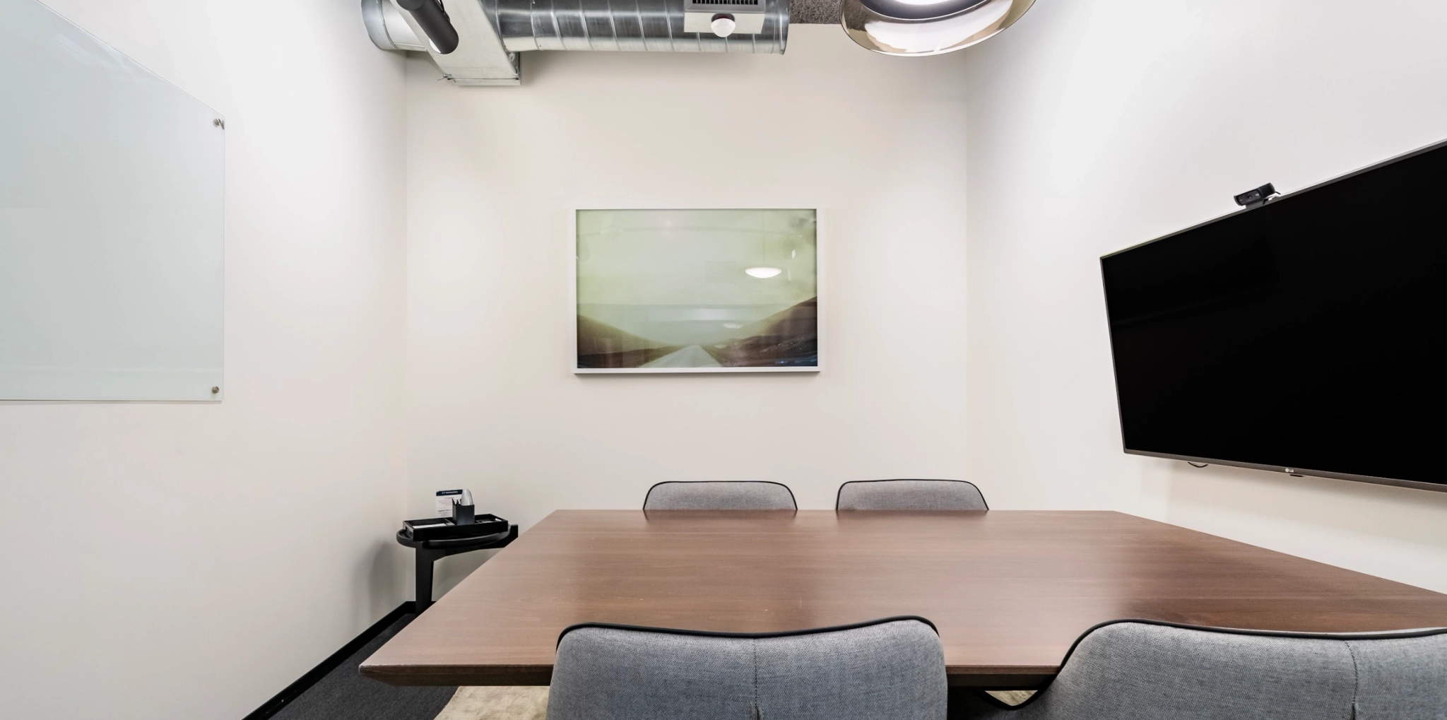 Un espace de coworking chaleureux comprenant une petite salle de réunion avec une table rectangulaire en bois, quatre chaises, un grand écran mural, un tableau blanc et une œuvre d'art abstraite.