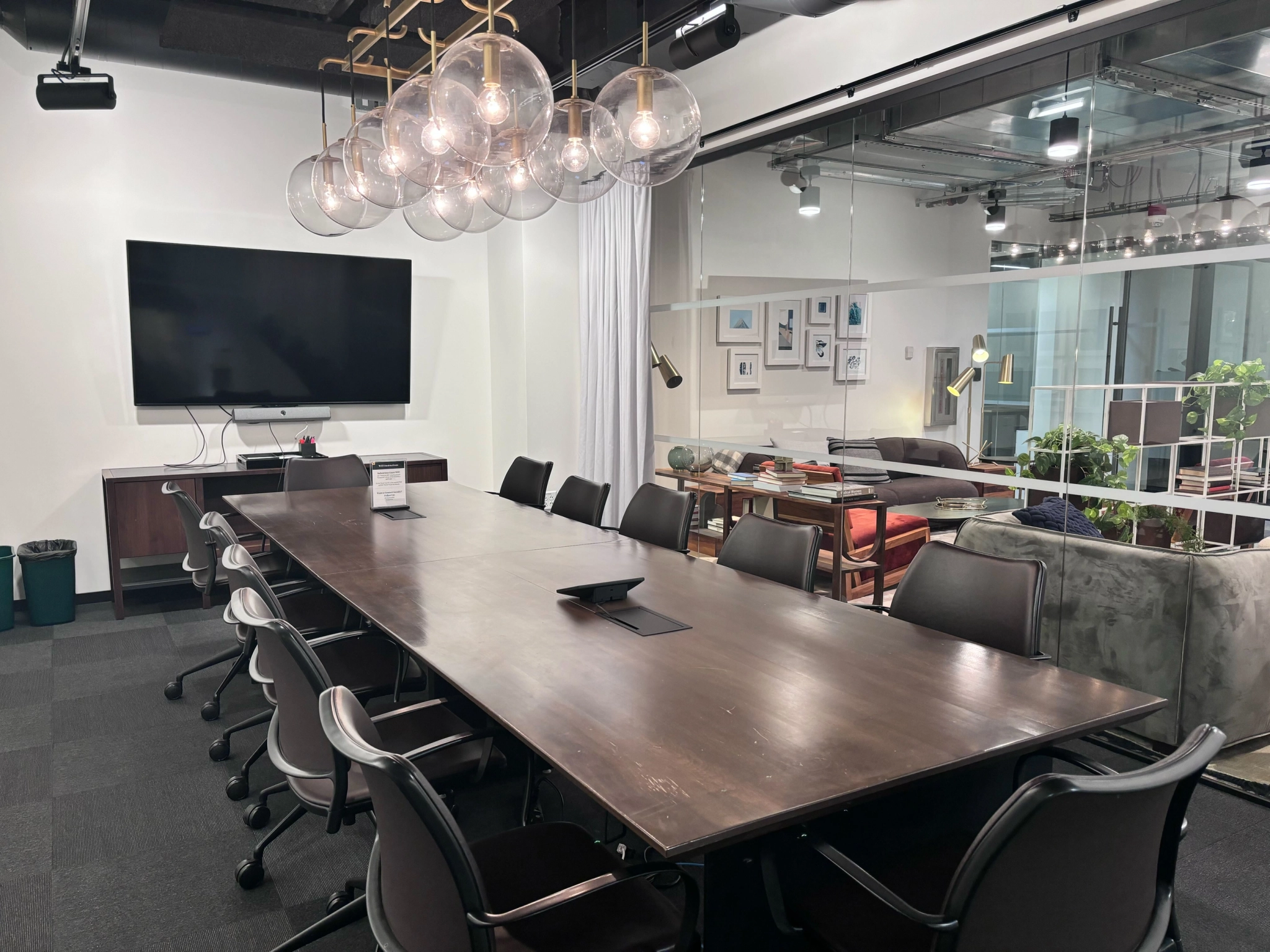 A modern office meeting room features a long wooden table, black chairs, a wall-mounted TV, and a glass partition. Hanging globe lights and framed pictures adorn this versatile workspace.