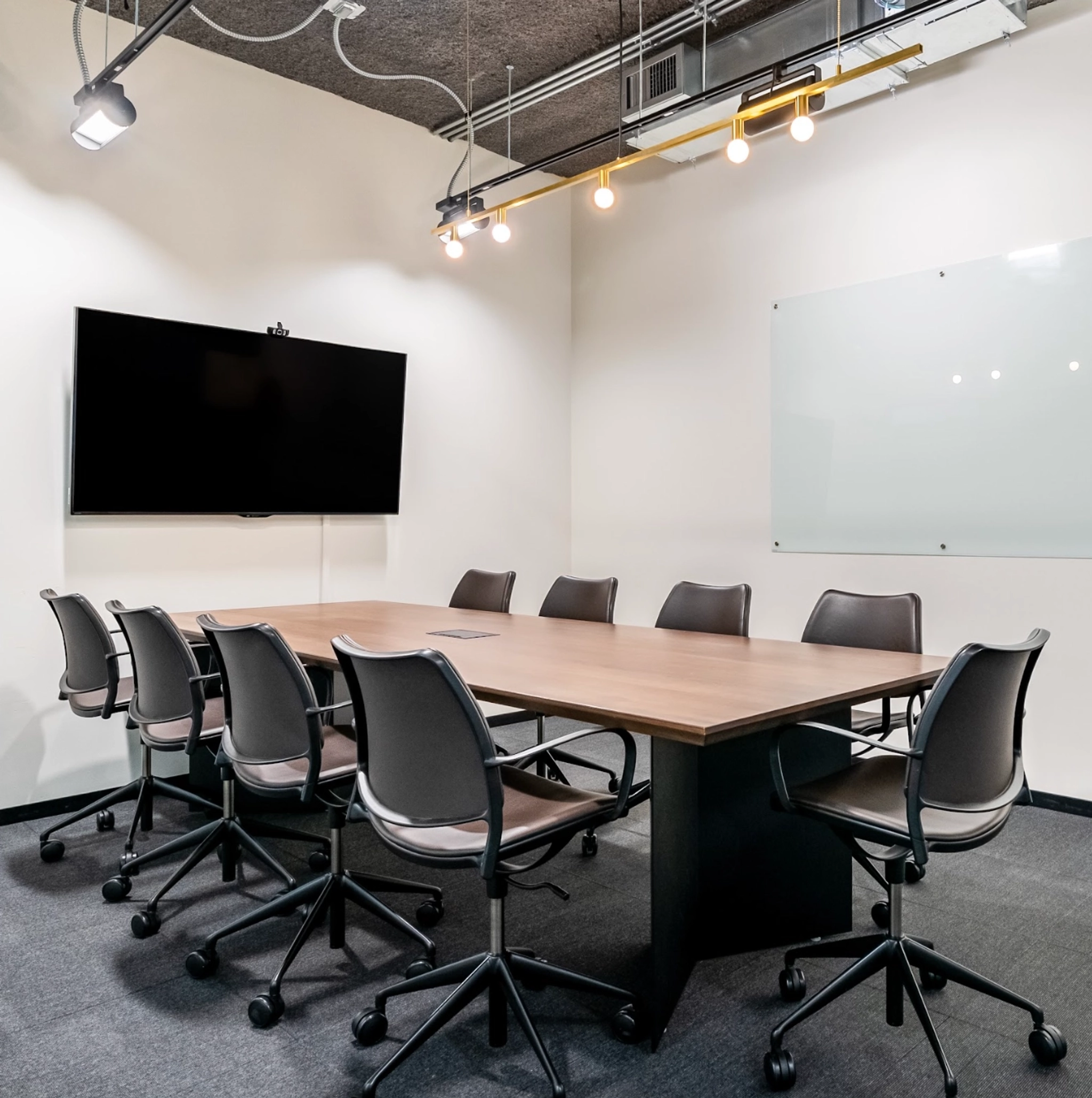 Une salle de réunion moderne dotée d'une table en bois, de huit chaises, d'un écran mural et d'un tableau blanc vierge. L'espace dispose d'un plafond exposé avec un éclairage industriel, parfait pour tout environnement de coworking.