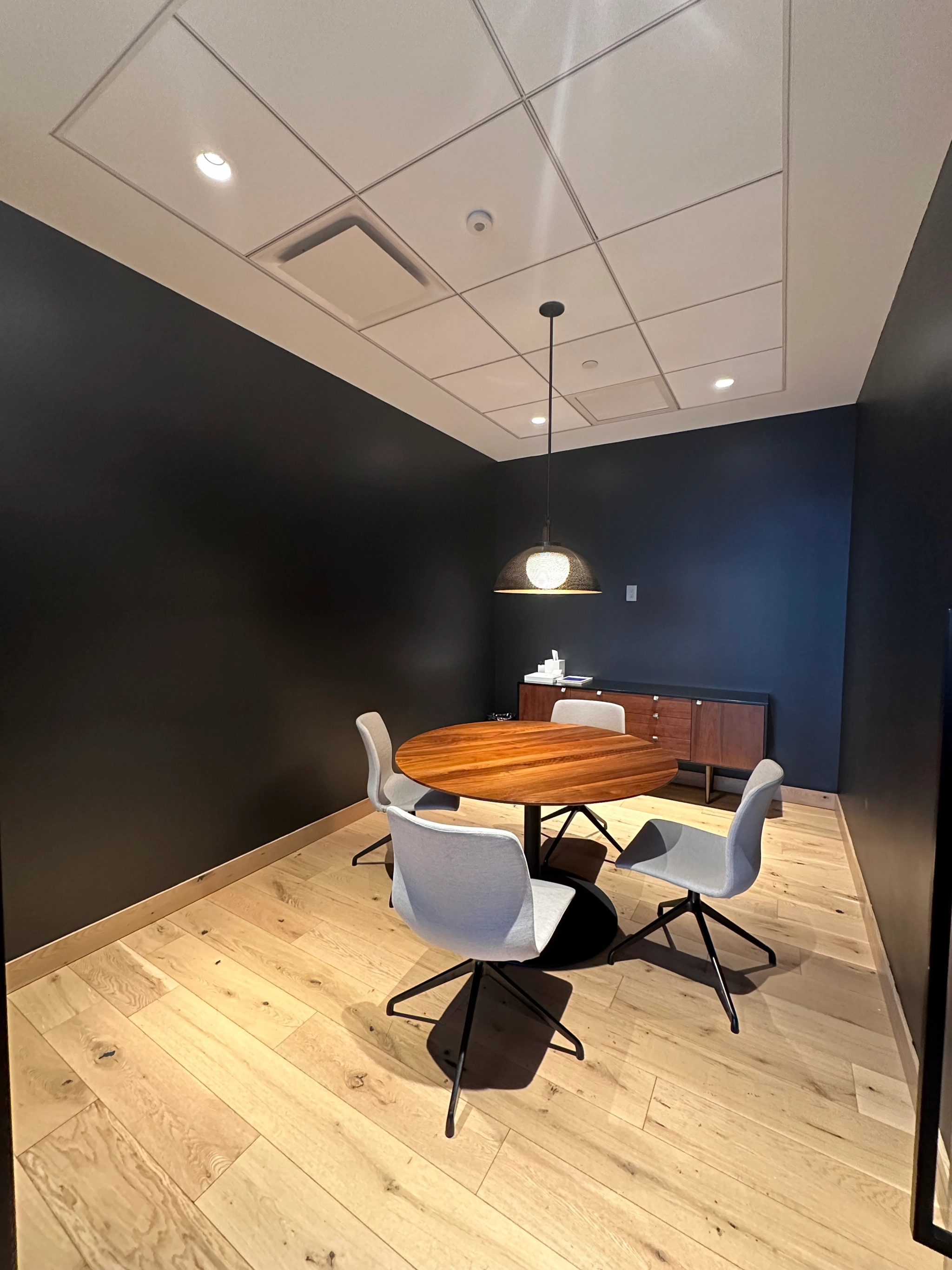 A cozy workspace features a round wooden table with three chairs, perfectly illuminated by a ceiling light. The meeting room boasts dark walls contrasting with a light wood floor, complemented by a side cabinet for storage.