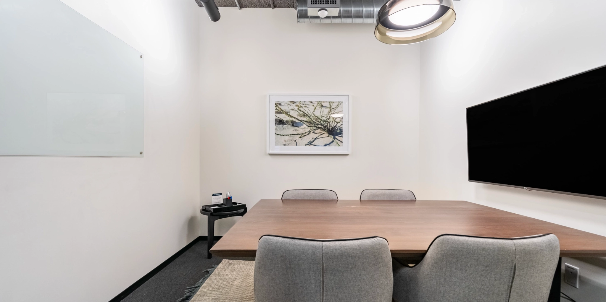 Une salle de réunion de bureau confortable comprend une table en bois, trois chaises grises, une télévision murale, un tableau blanc et des œuvres d'art encadrées sur le mur, parfaites pour les séances de coworking.