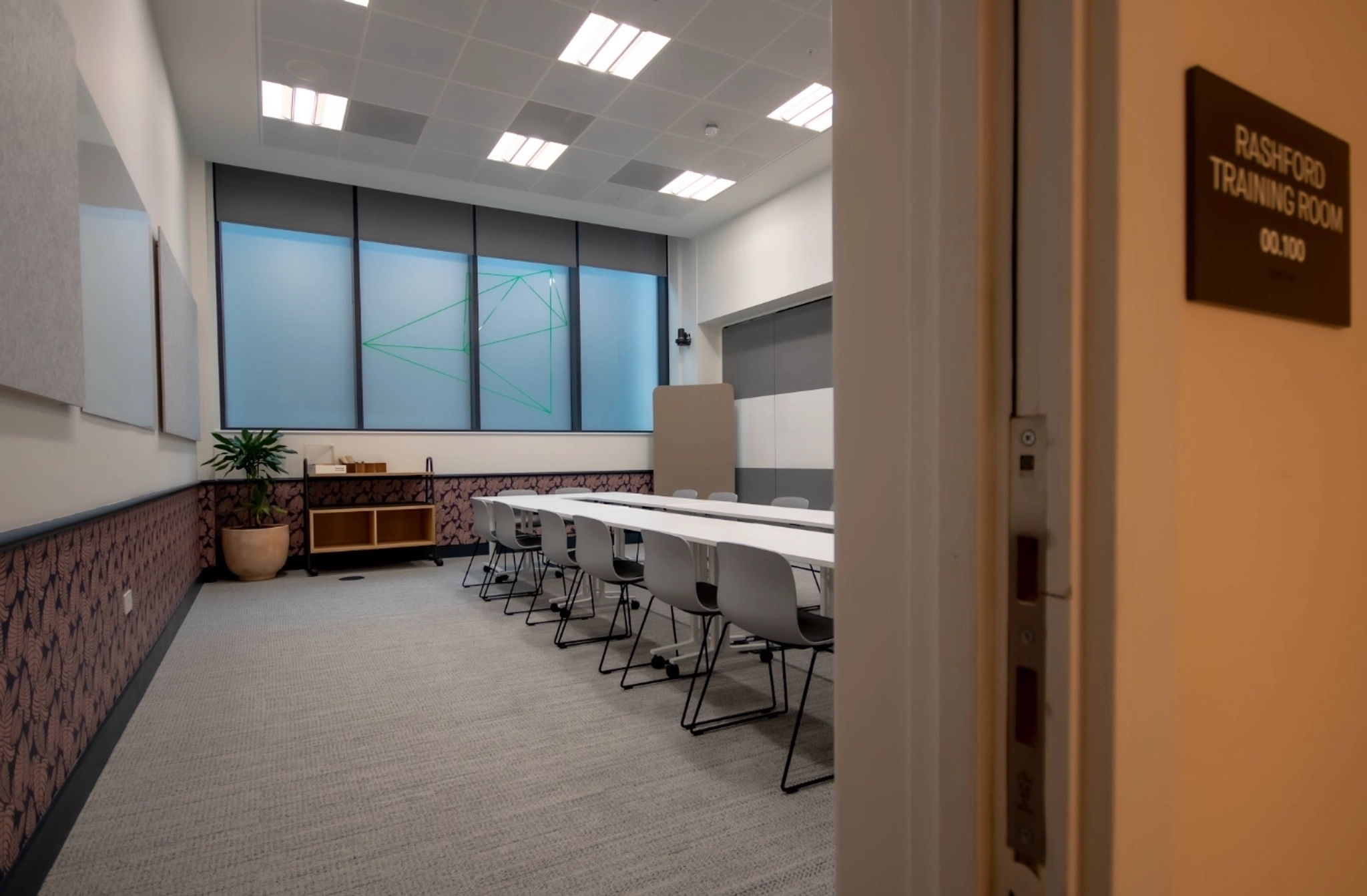 A modern Manchester meeting room with a long table, gray chairs, wall panels, a large plant, sideboard, and a sign reading "Rashford Training Room 00.100" by the door—an inviting workspace for any gathering.