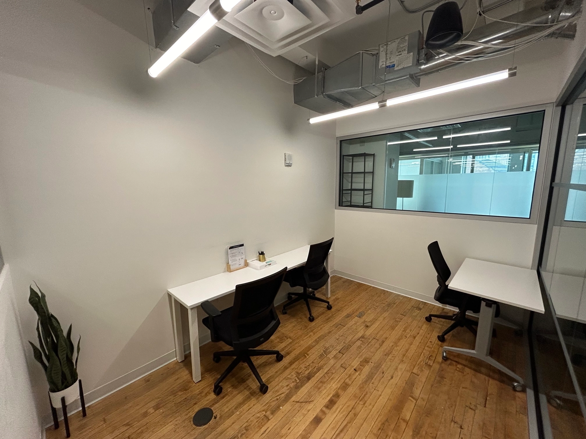 Un petit espace de travail moderne avec trois bureaux, trois chaises noires, une plante en pot, un sol en bois et une fenêtre donnant sur un autre bureau.