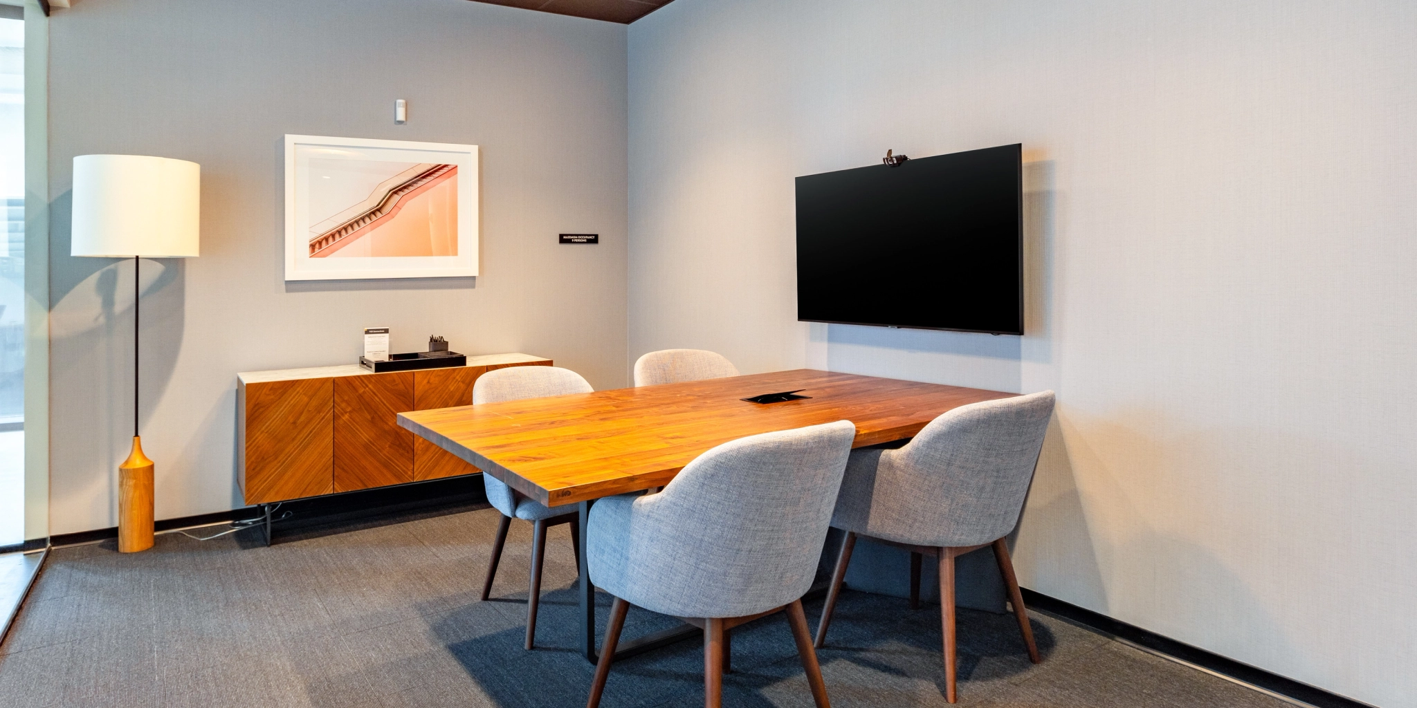 A small meeting room with a wooden table, four chairs, a wall-mounted TV, a framed picture, and a lamp. The workspace is complemented by a cabinet against the gray wall.