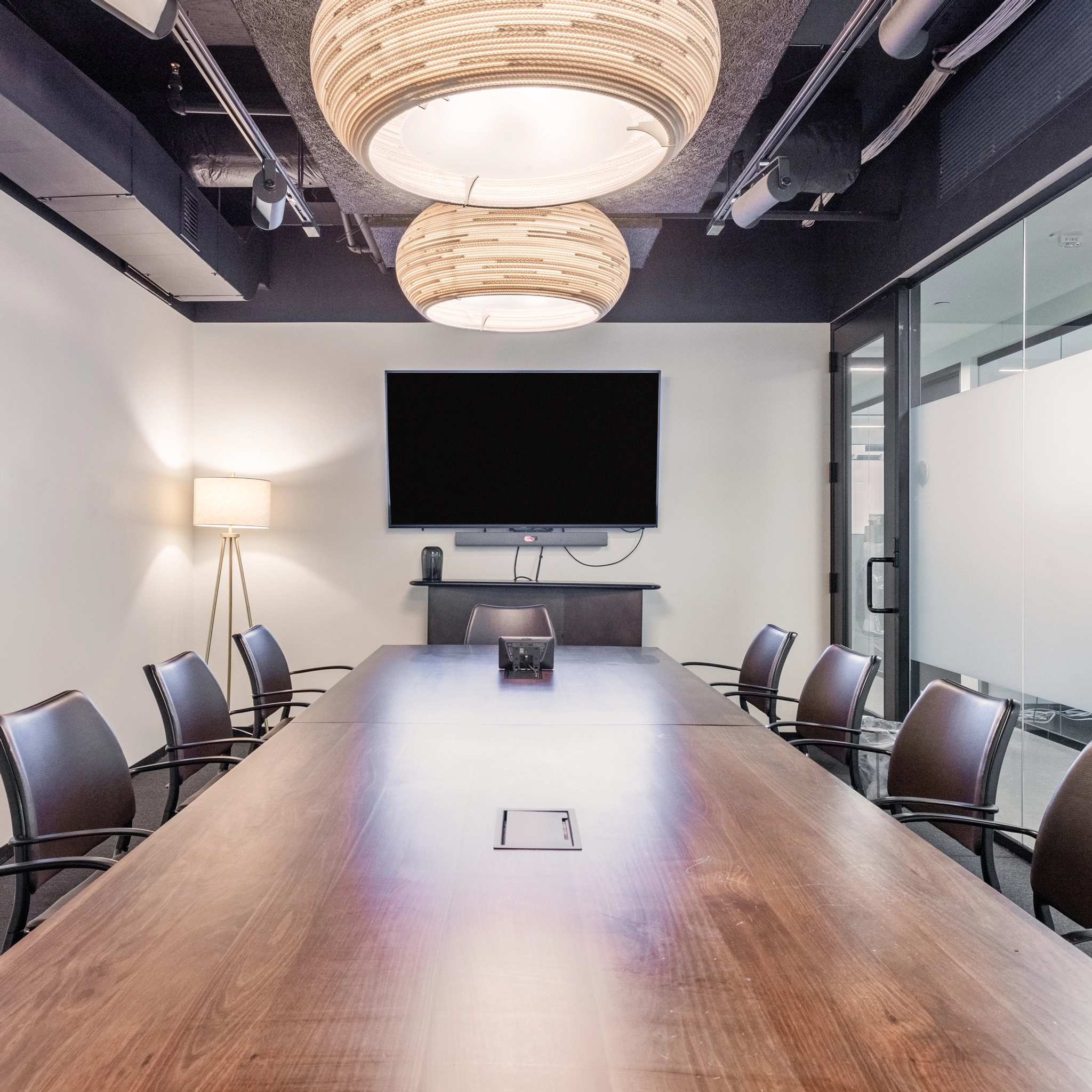 A modern coworking conference room with a long wooden table, black chairs, a wall-mounted TV, and two circular light fixtures.