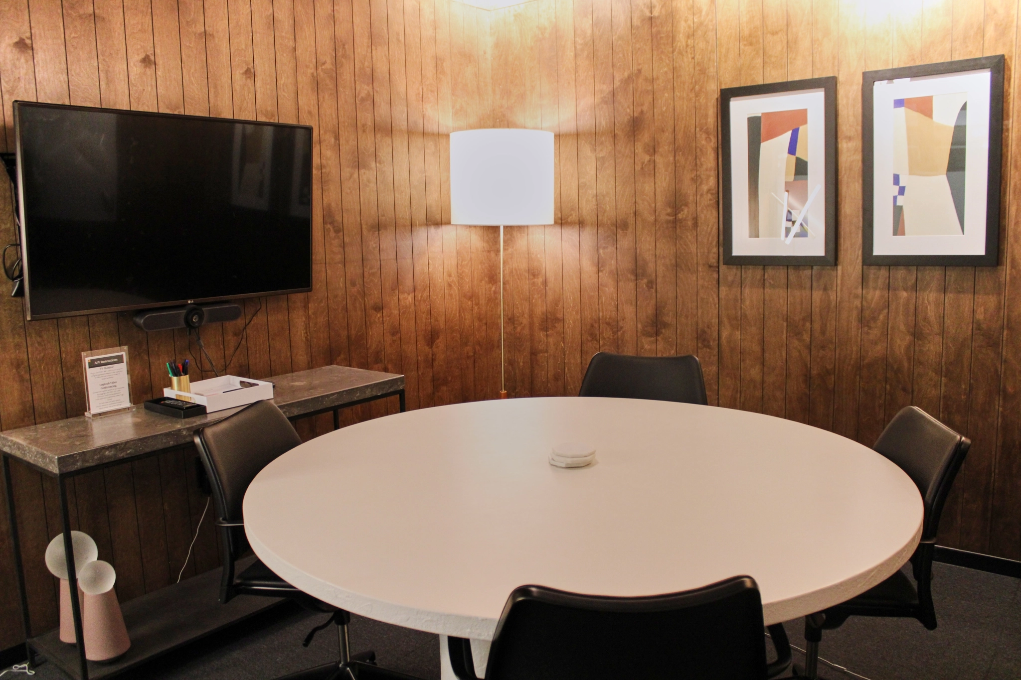 A small coworking meeting room in Atlanta with a round white table, four chairs, a wall-mounted TV, wall art, a lit floor lamp, and a console table holding stationery and a small device. The wood-paneled walls add a touch of warmth to this efficient office space.