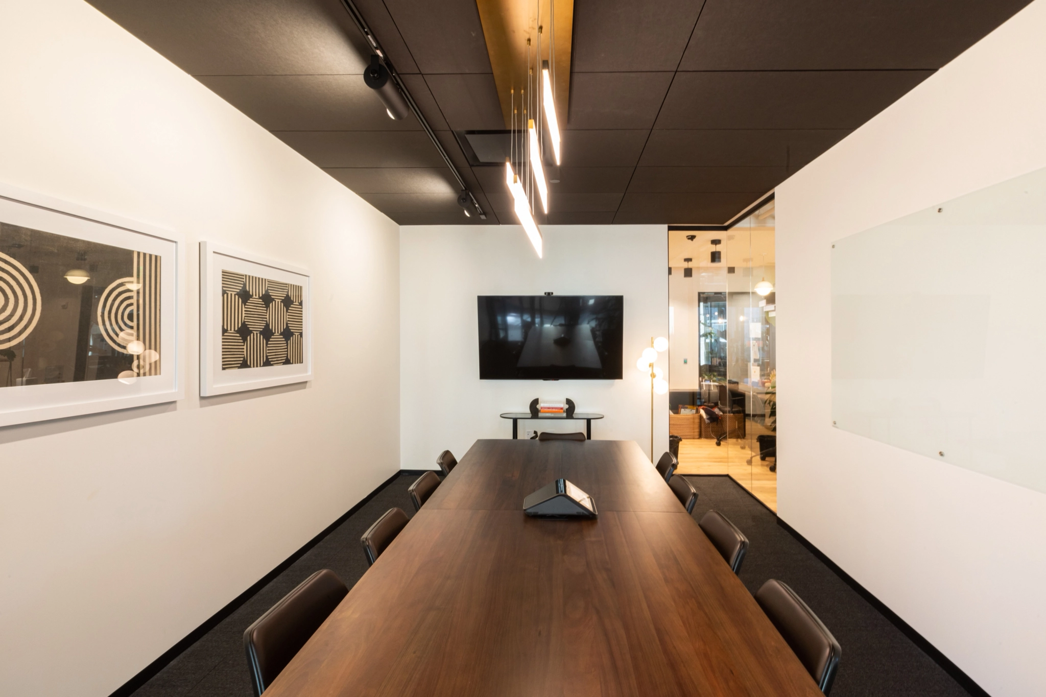 A modern coworking space in Arlington features a spacious meeting room with a long wooden table, eight chairs, a large wall-mounted screen, abstract artwork, and a glass whiteboard. Ceiling lights illuminate the room.