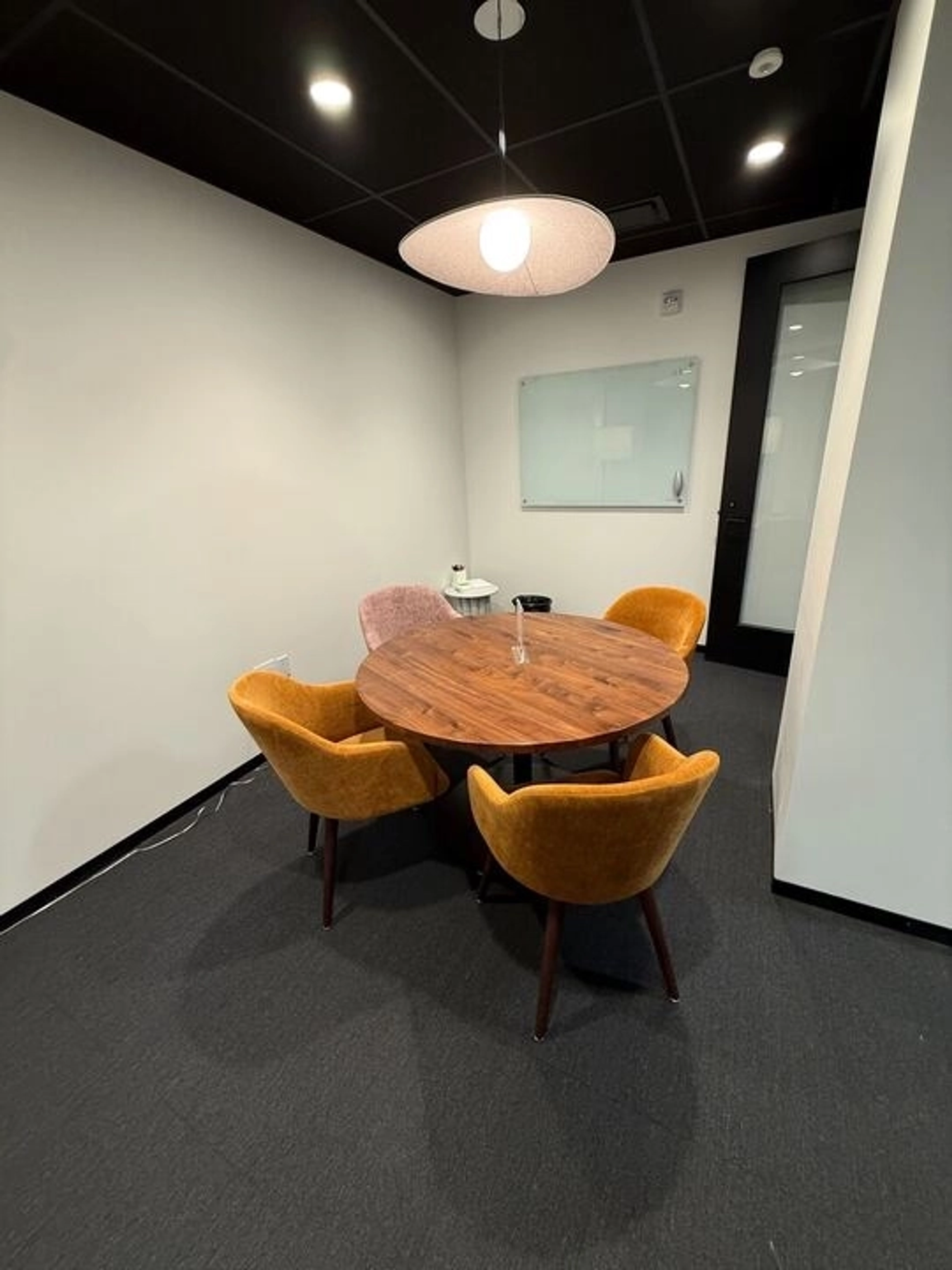 Une petite salle de réunion à Washington avec une table ronde en bois, quatre chaises rembourrées, un plafonnier, un tableau blanc en verre sur le mur et une porte fermée - parfait pour un espace de travail ciblé.