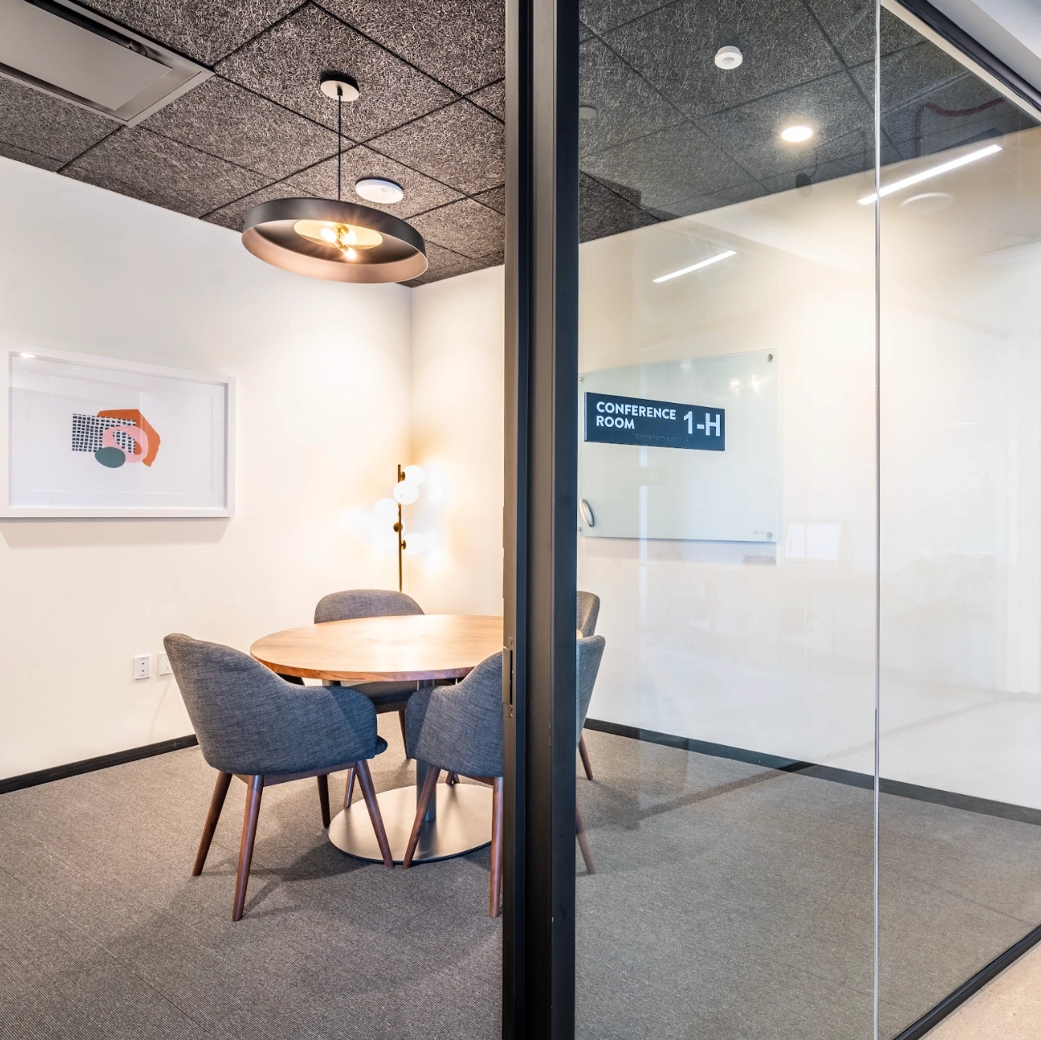 Image of a small meeting room with a round wooden table and four chairs. The room has a glass door with a sign that reads "Conference Room 1-H" and a modern ceiling light fixture, perfect for coworking or small office gatherings.