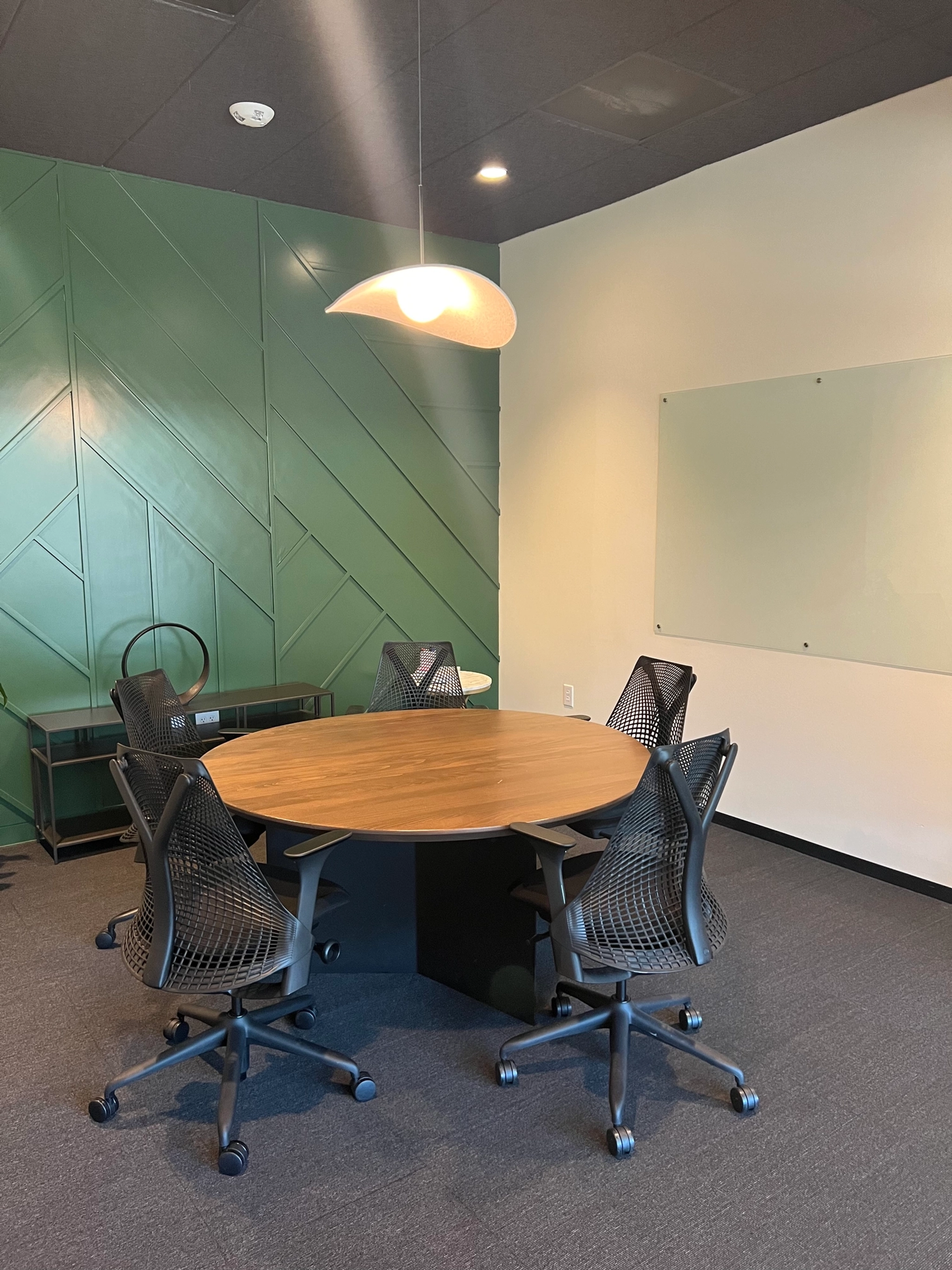 A modern coworking space featuring a round wooden table, five black mesh chairs, a green accent wall, a whiteboard, and a hanging light fixture.