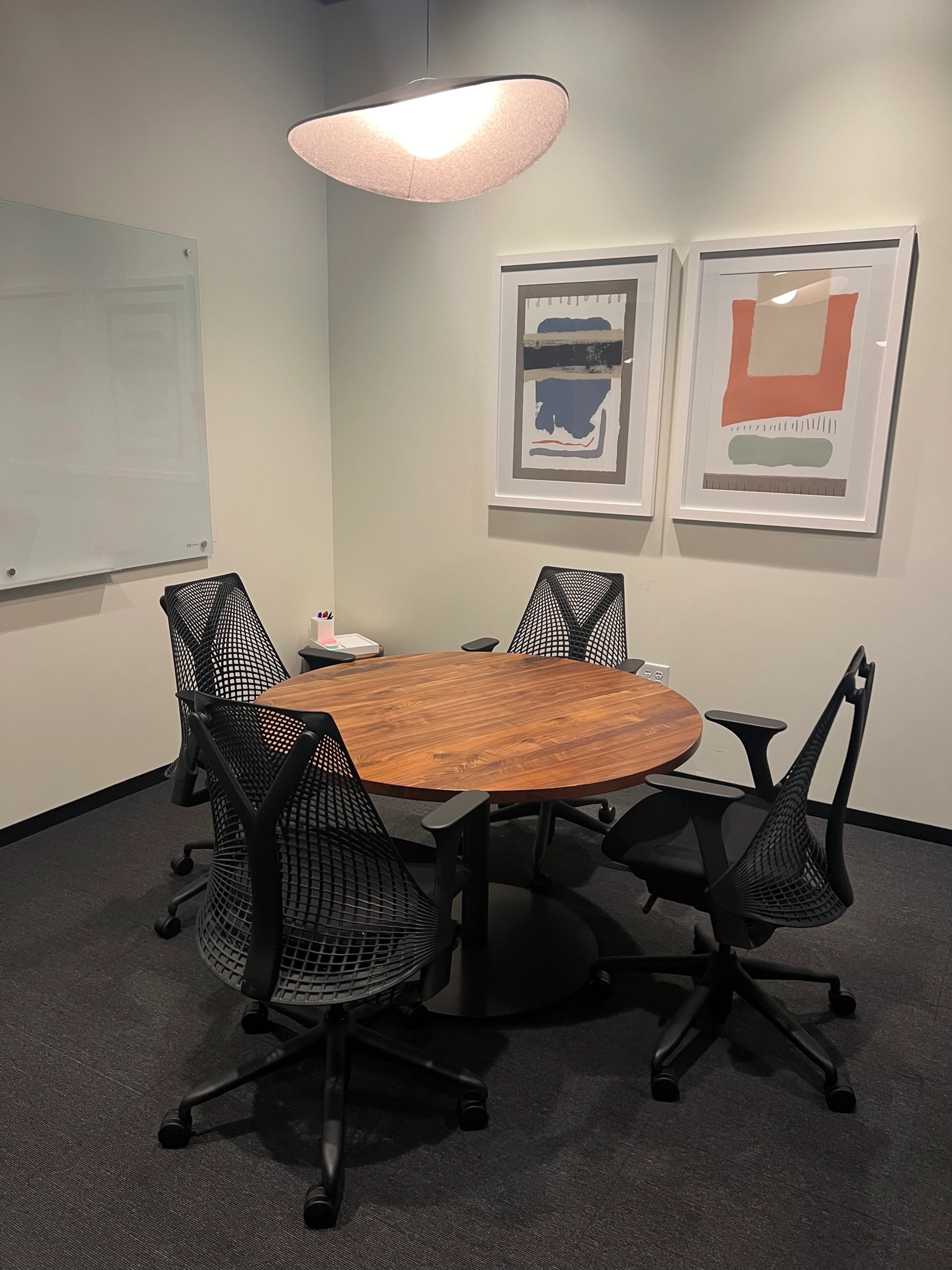 Une petite salle de conférence dans notre bureau de Dallas comprend une table ronde en bois entourée de quatre chaises en filet noir, un tableau blanc au mur et deux œuvres d'art abstraites accrochées au-dessus de l'espace de travail.