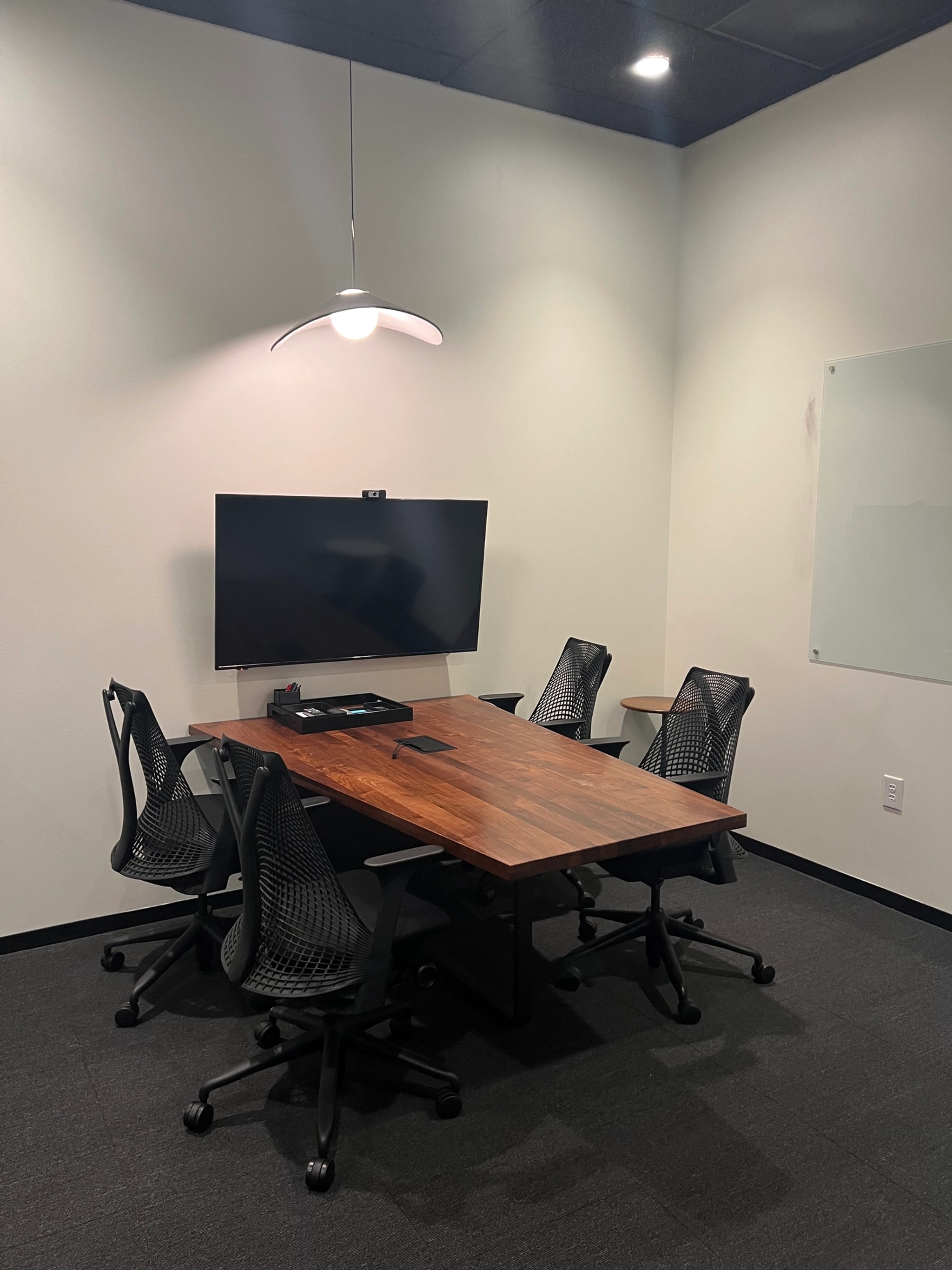 A small conference room in the coworking space features a wooden table, four mesh chairs, a wall-mounted screen, and a light fixture overhead. A whiteboard is mounted on the right wall.