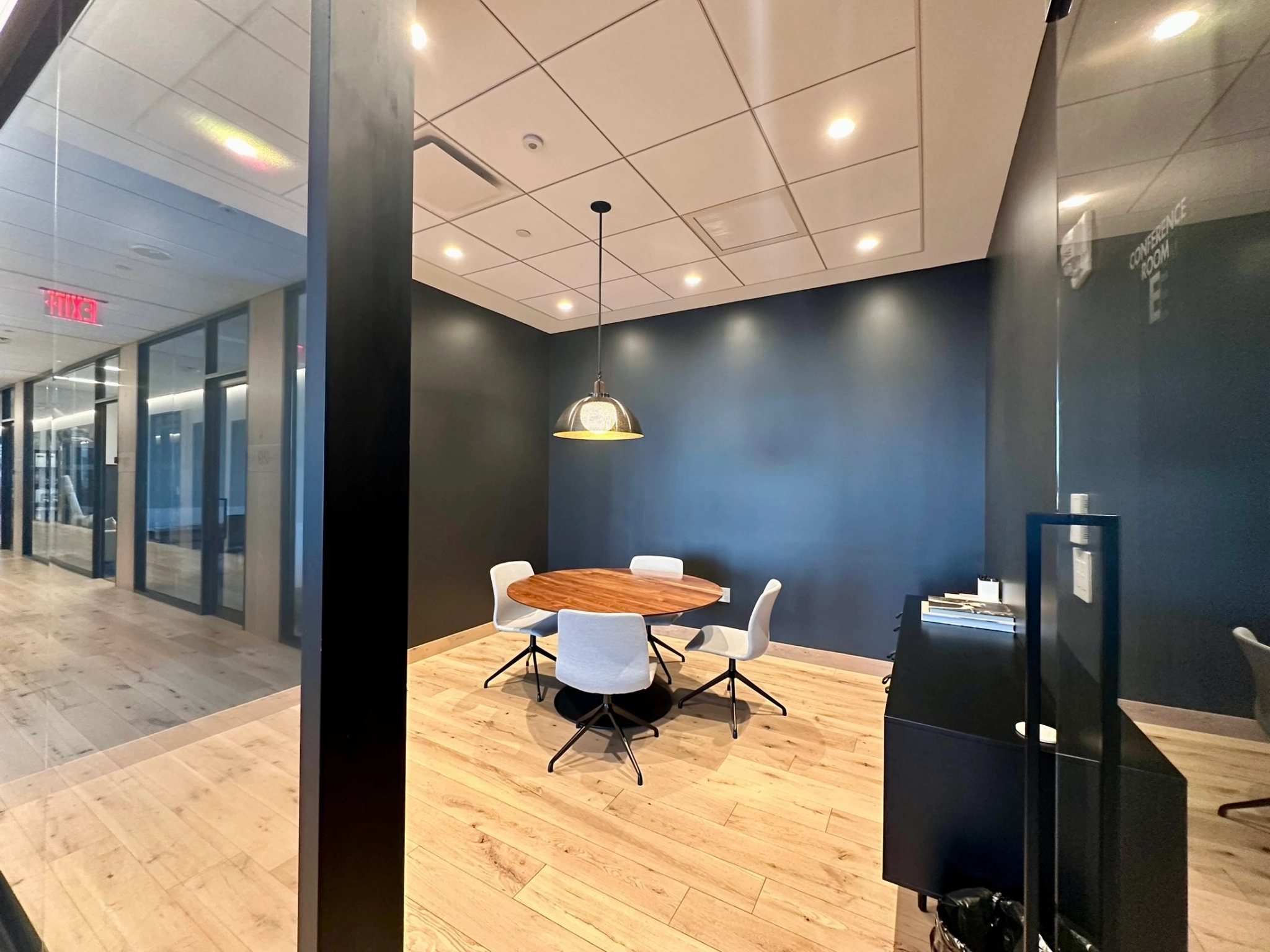 A modern coworking space features a round wooden table, four chairs, a ceiling light, and wood flooring, making it the perfect meeting room for collaboration.